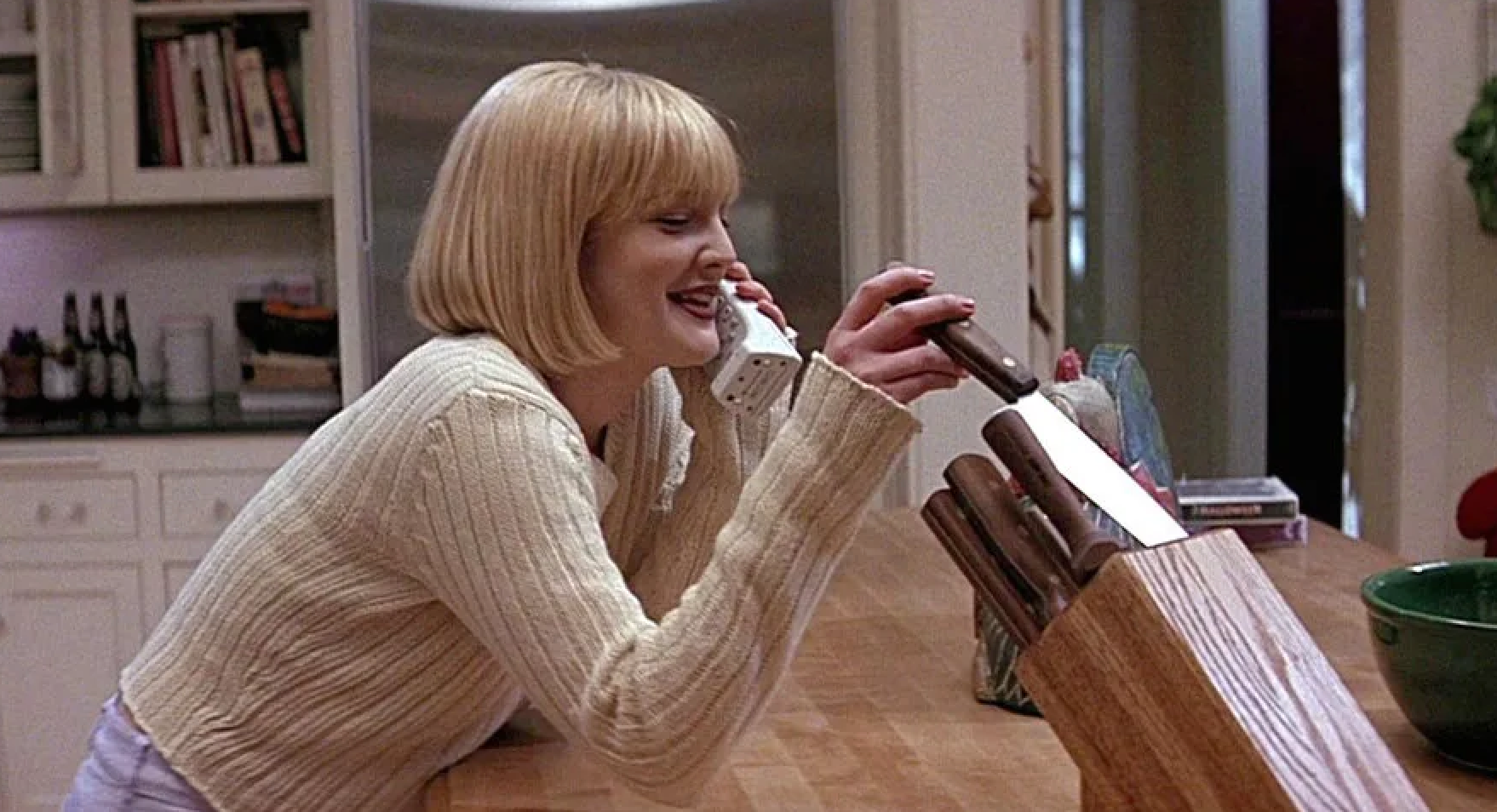 Person in a knit sweater holds a phone and examines a knife block in a kitchen, reminiscent of a suspenseful movie scene