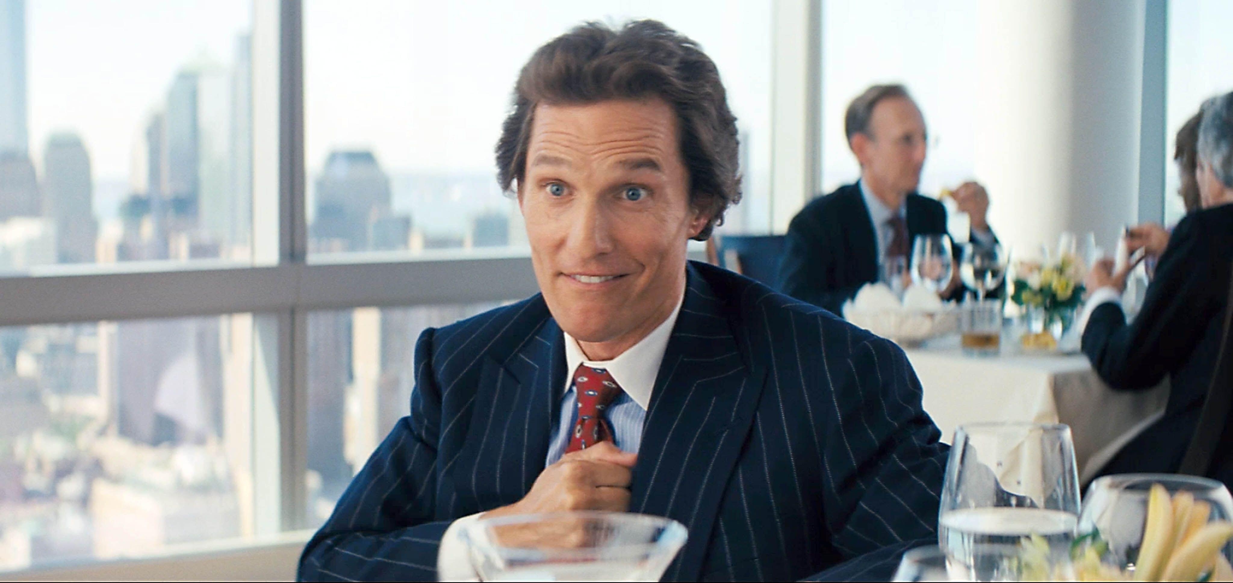 Person in a pinstripe suit enthusiastically gestures while sitting at a restaurant table with cityscape in the background