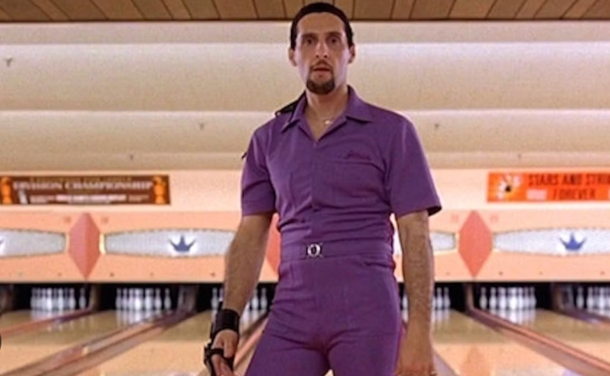 Person in a bowling alley wearing a purple jumpsuit, holding a bowling ball