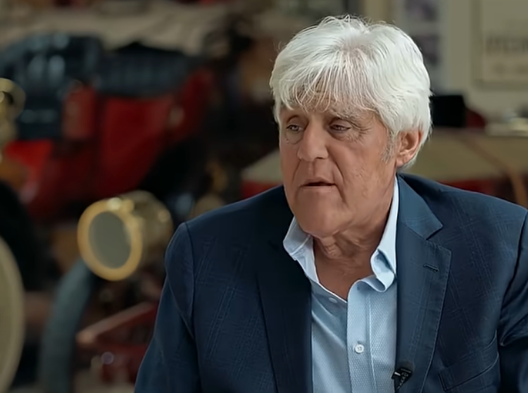 Person in a blue suit jacket and open-collar shirt speaking indoors
