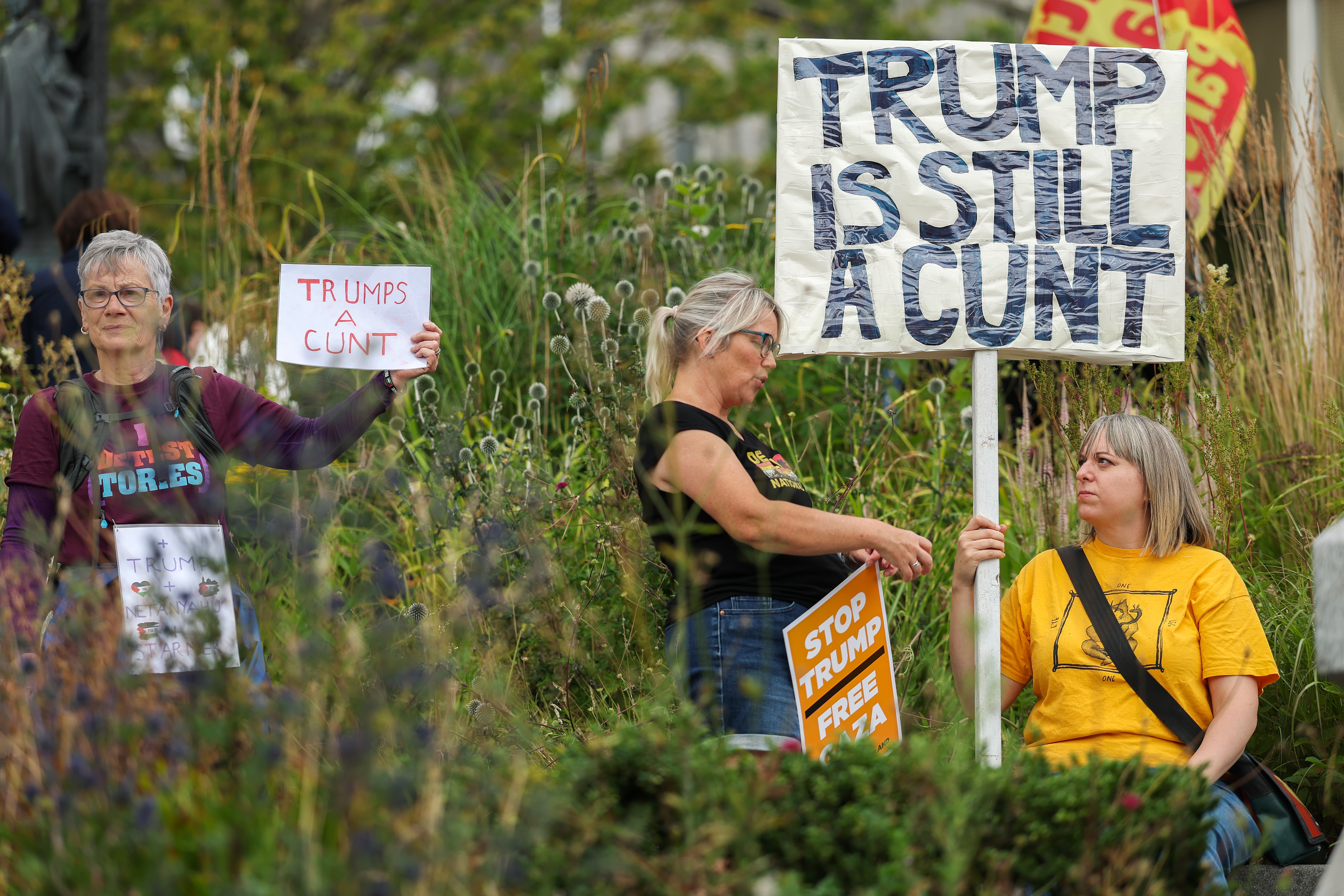 Three protesters hold signs with anti-Trump messages in a garden setting