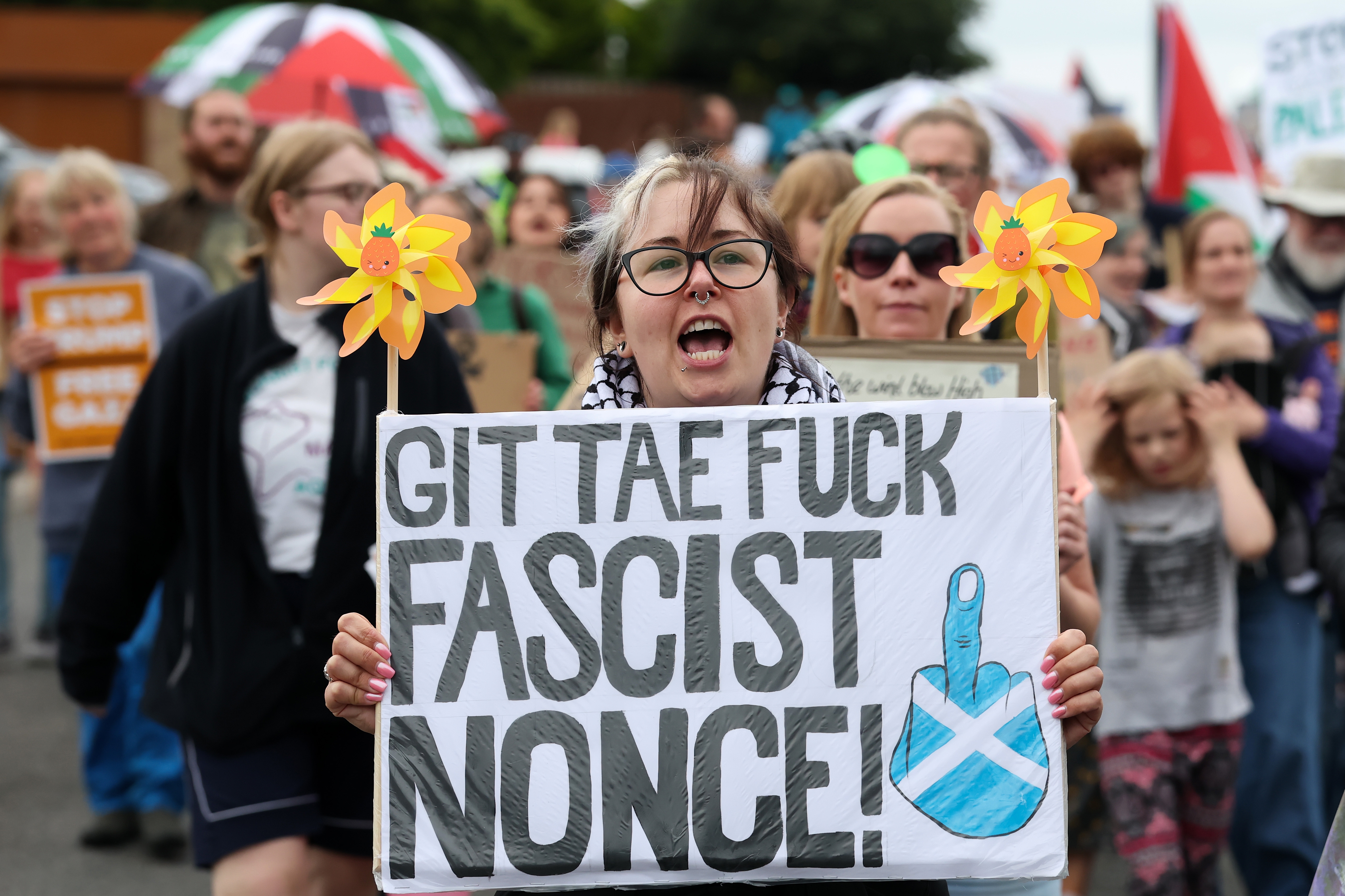 Person holding protest sign with bold text and Scottish flag, surrounded by a crowd at a public demonstration