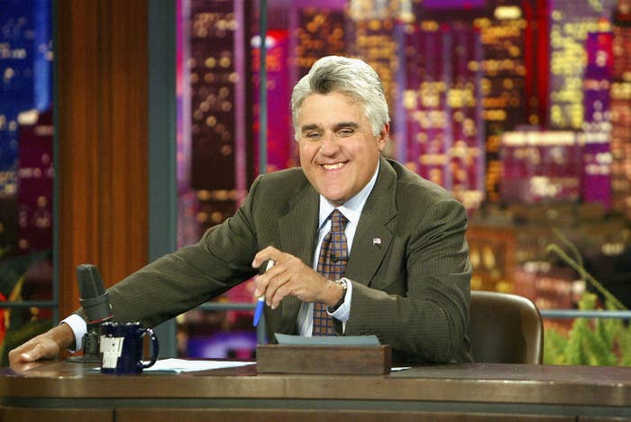 A person in a suit sits at a late-night talk show desk, smiling, with a cityscape backdrop, holding a pen