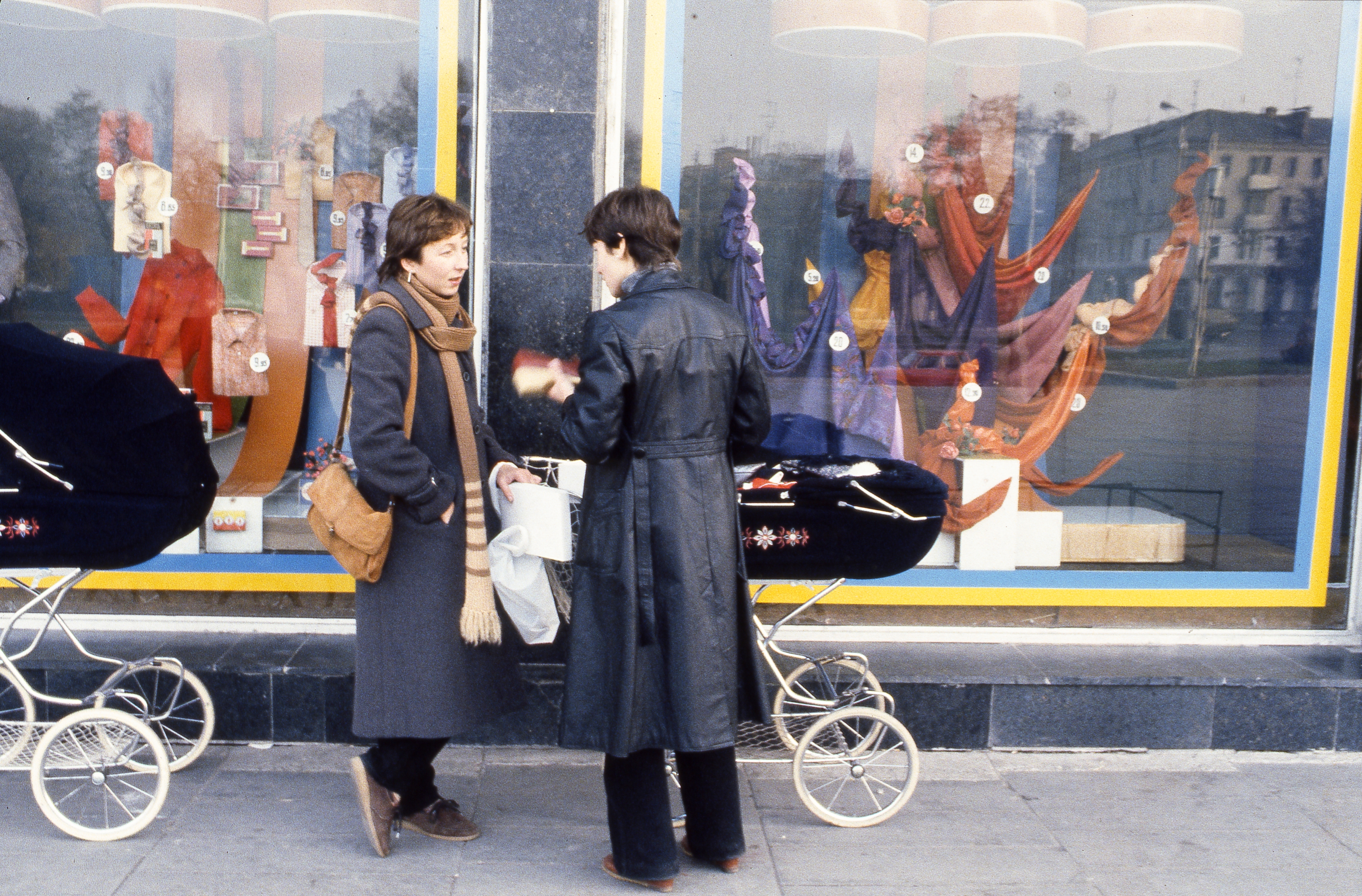 Two people stand outside a shop window with mannequins, talking near baby carriages. Warm coats and scarves suggest a colder climate