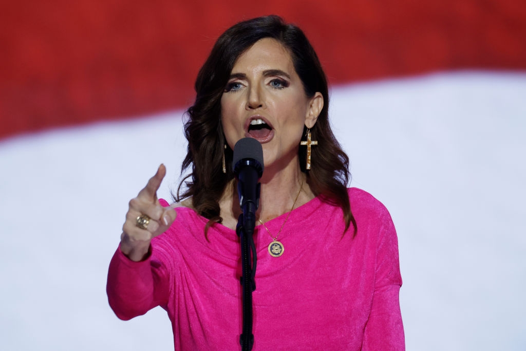 A person speaks passionately at a microphone, wearing a pink top and cross earrings, set against a bold background