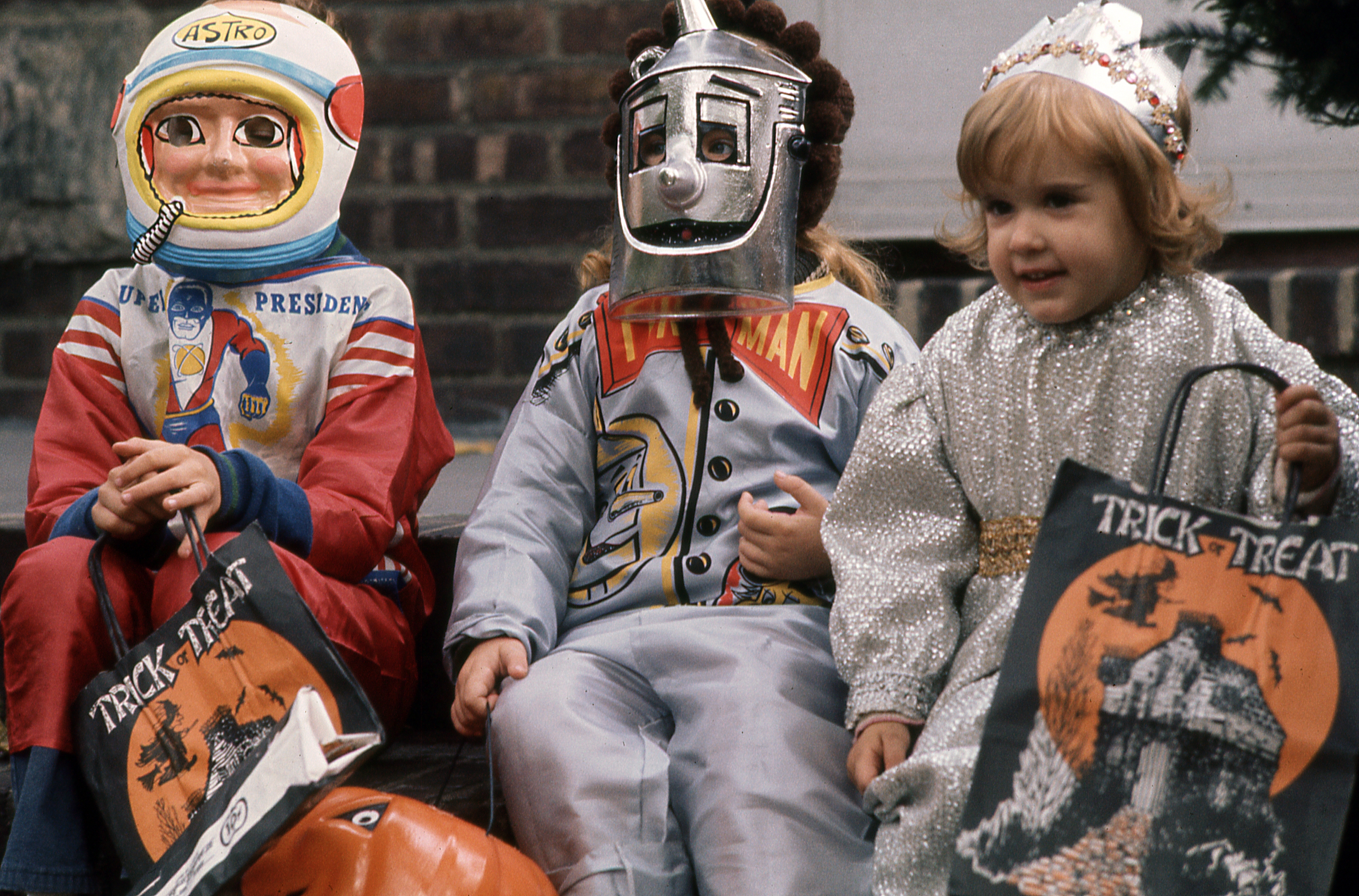 Three children in vintage Halloween costumes: astronaut, robot, and a princess. They sit on steps holding trick-or-treat bags with Halloween designs