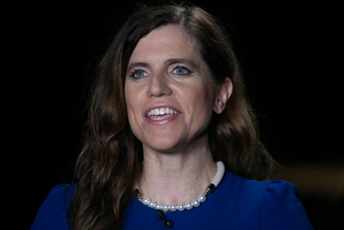 A person with long hair, wearing a blue top and a pearl necklace, smiling in a dimly lit setting