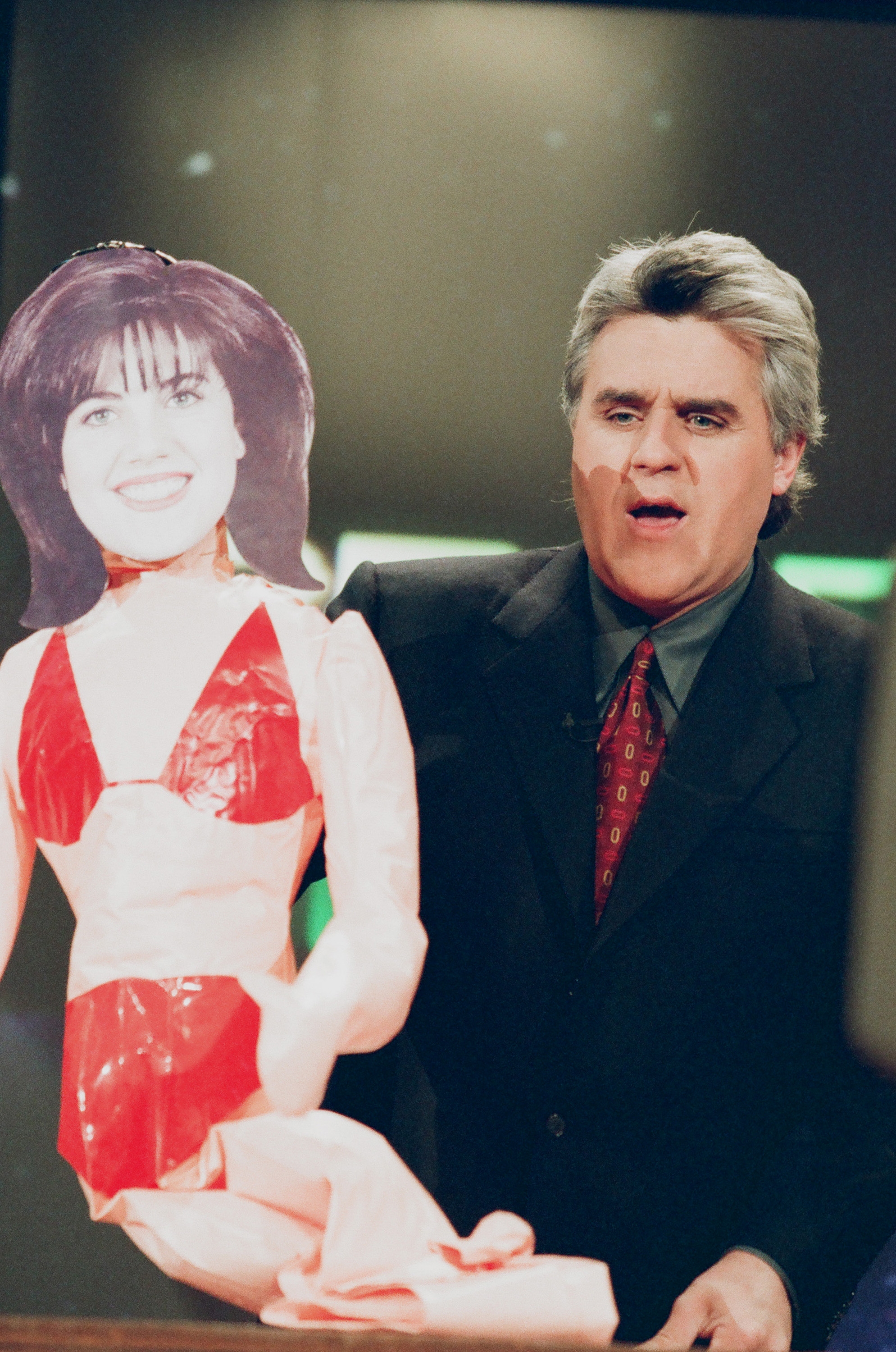 A man in a suit holds a humorous cardboard cutout of a woman on a talk show set, adding a playful element to the scene