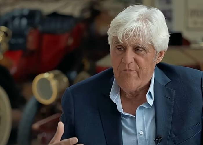 Man in blazer talks with hand gesture, background of vintage cars, suggesting an interview or discussion setting