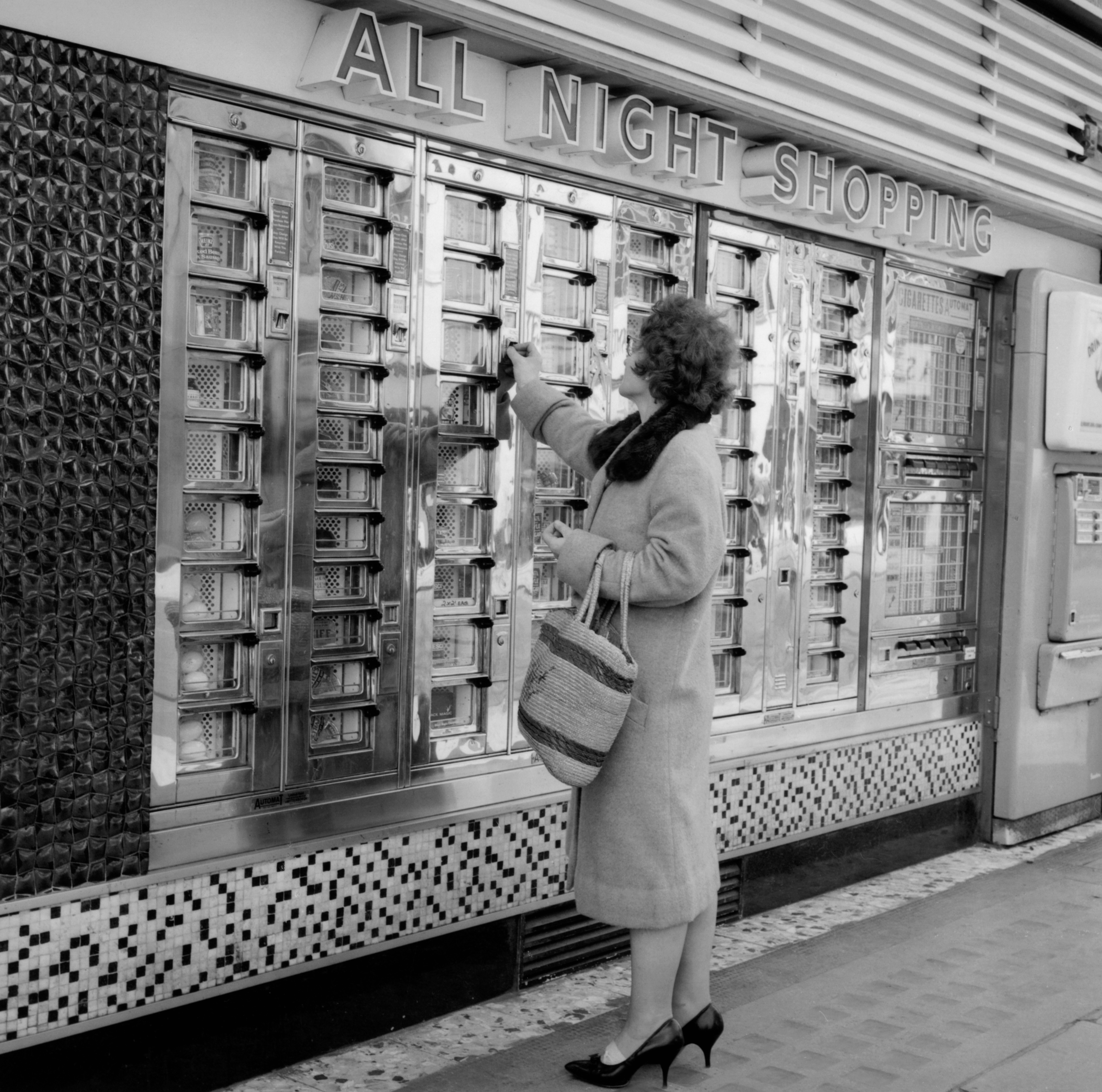 A woman in a coat and heels uses a vintage vending machine labeled "All Night Shopping" while holding a handbag