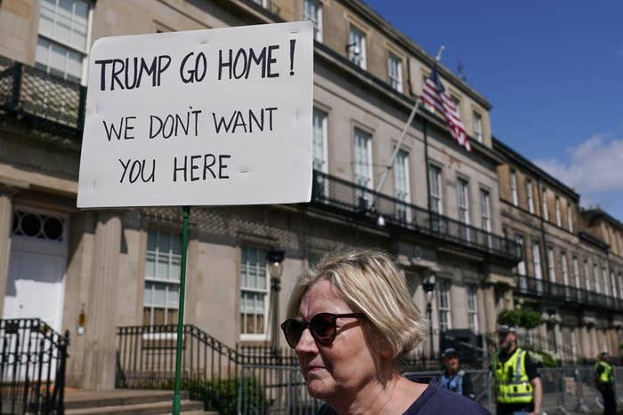 A woman stands at a protest holding a sign reading &quot;TRUMP GO HOME! WE DON&#x27;T WANT YOU HERE&quot; outside a historic building with an American flag