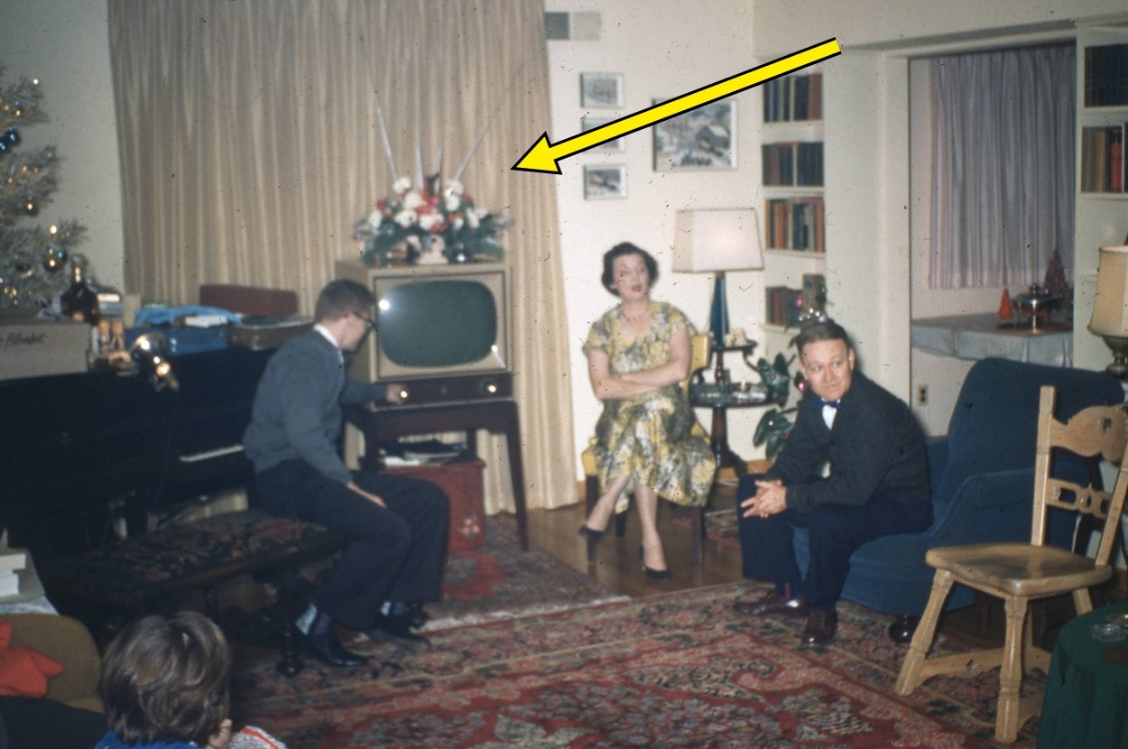 Three people sit in a living room with vintage decor. A TV has a flower arrangement on top. Arrow points to it