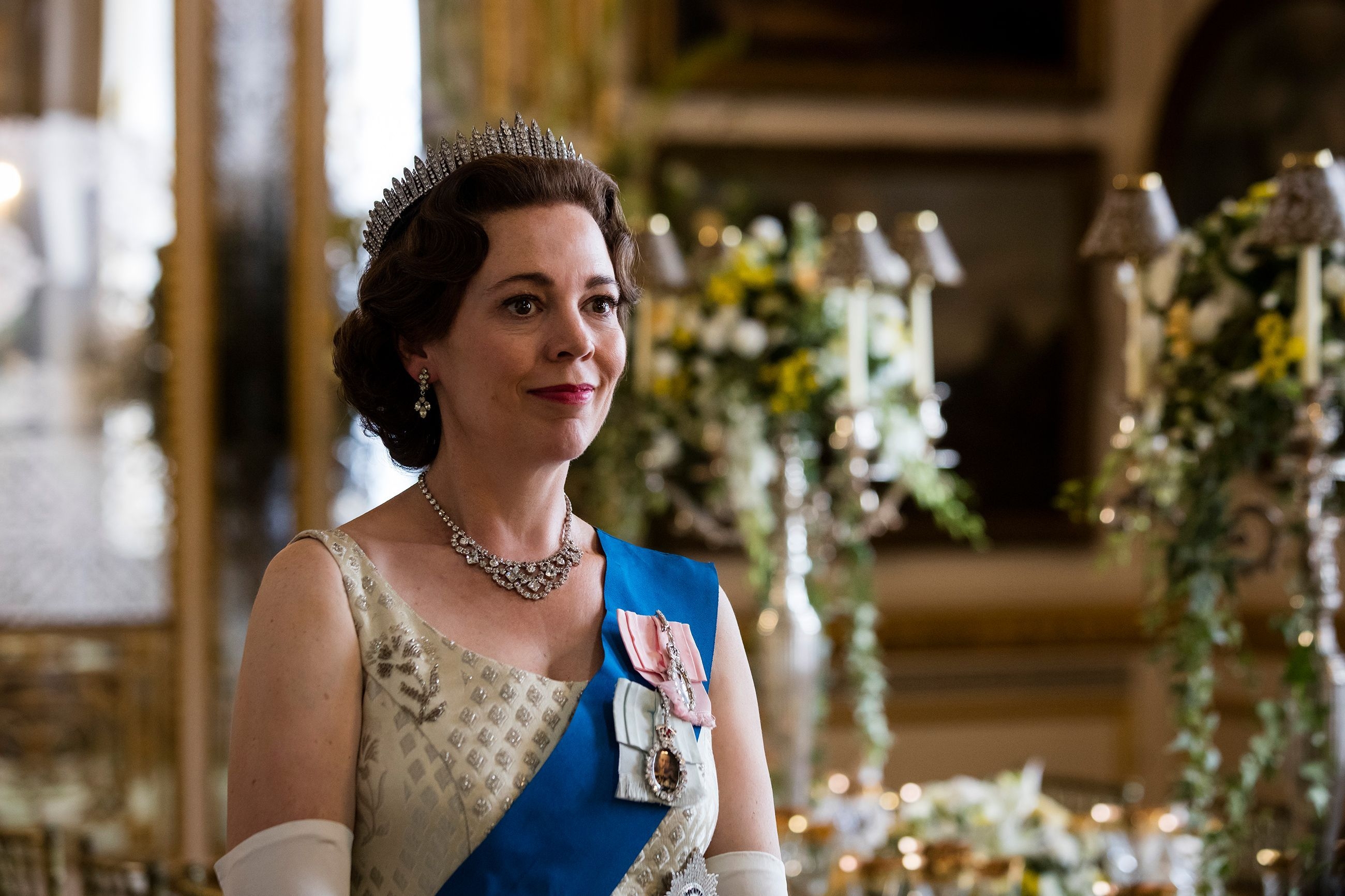 Persona con vestido de gala y tiara en un entorno real, sonriendo, posiblemente en una escena de TV o película de época