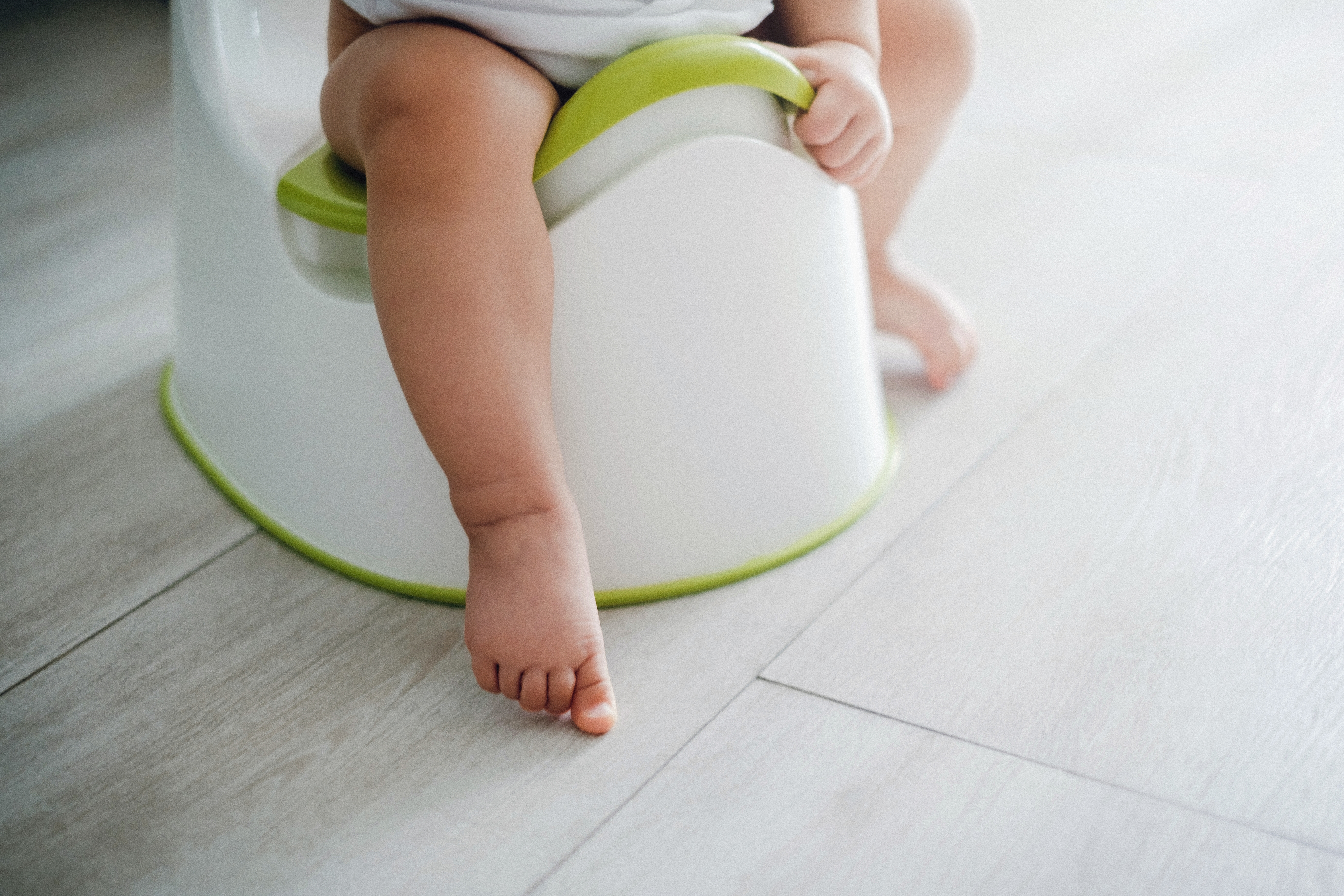 A toddler's legs are visible while they sit on a potty training seat on a wooden floor