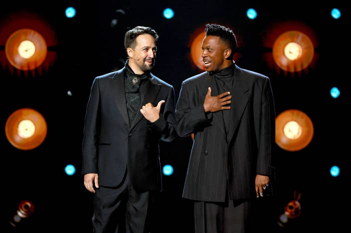Two people wearing stylish black suits stand on stage, one gesturing to the other. Circular lights create a dramatic background effect