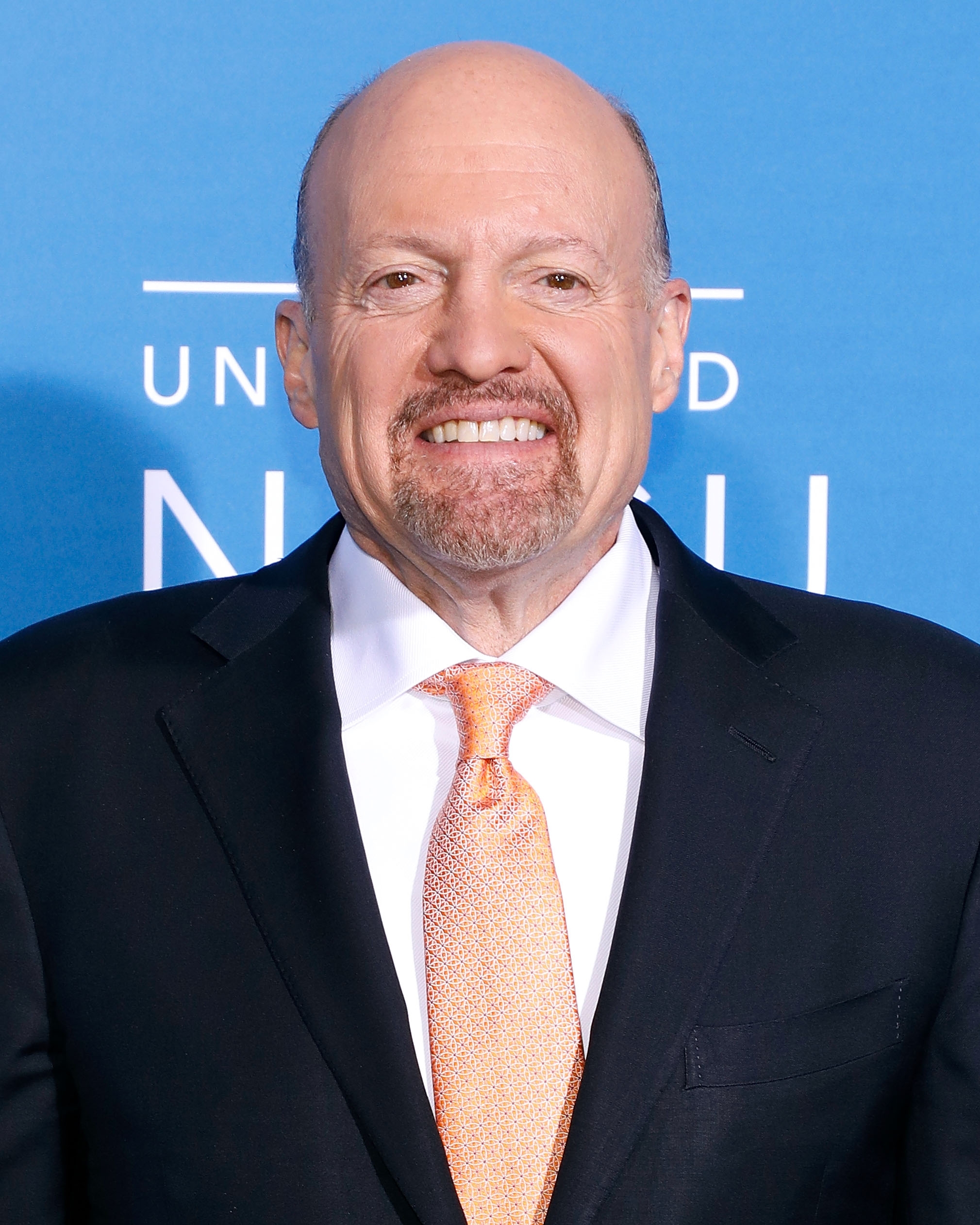 A person in a suit with a patterned tie is smiling at an event against a blue backdrop