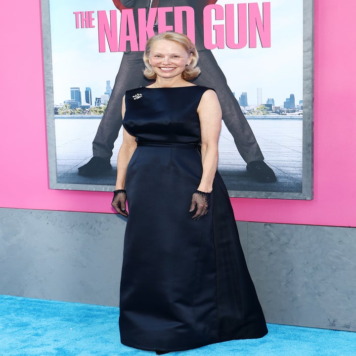 A person poses on the red carpet in a sleek black gown with a brooch and mesh gloves, with "The Naked Gun" poster in the background