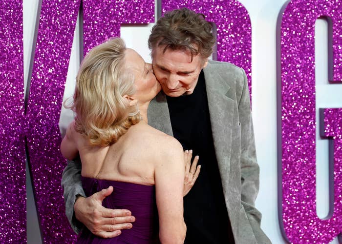 Two people embrace on the red carpet. One wears a stylish off-shoulder gown, the other a casual blazer. Background features large sparkly letters