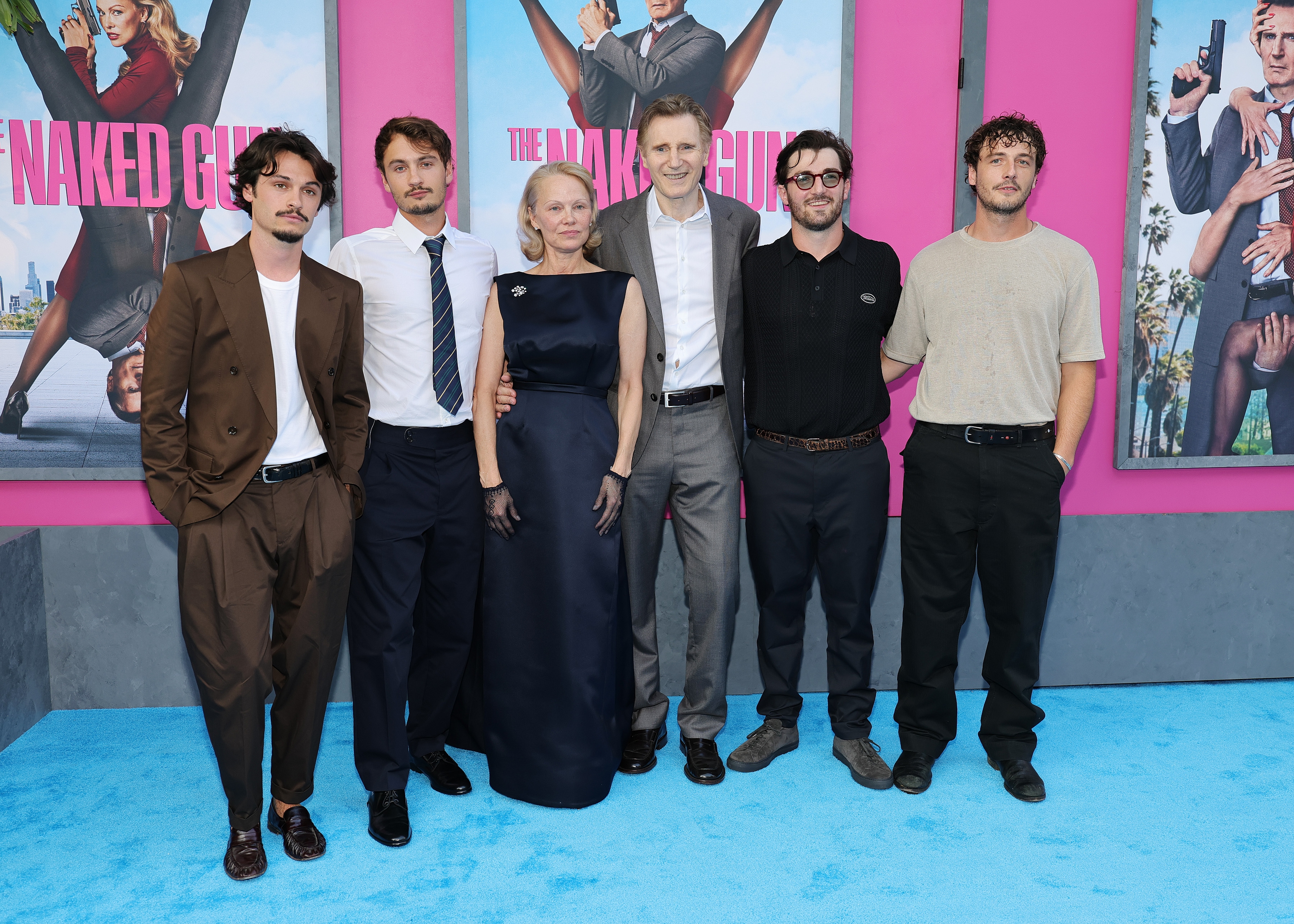 Group of people standing on a blue carpet, some in formal attire. Background has posters of "The Naked Gun."
