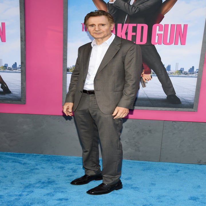 Person in a tailored suit stands on a blue carpet at a movie premiere. Movie poster visible in background
