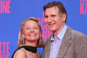 Two people pose together on a red carpet. One wears a sleek black dress, the other a light suit with a shirt, smiling for the cameras