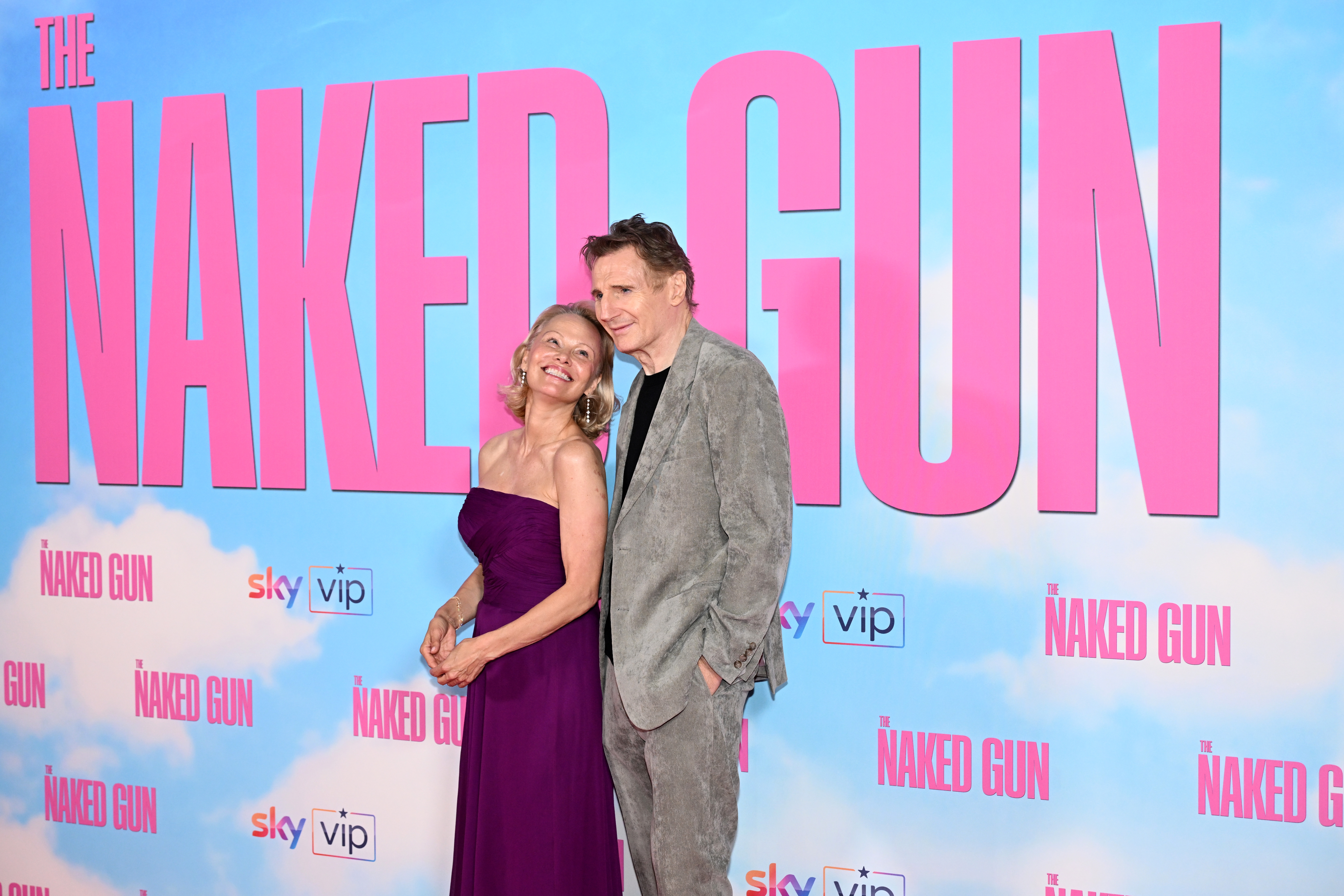 Two people pose on the red carpet at a "Naked Gun" event. One is in a formal purple dress, and the other in a gray suit