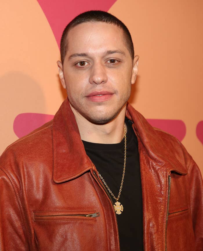 Pete Davidson in a leather jacket with a chain necklace poses for a photo against a geometric backdrop