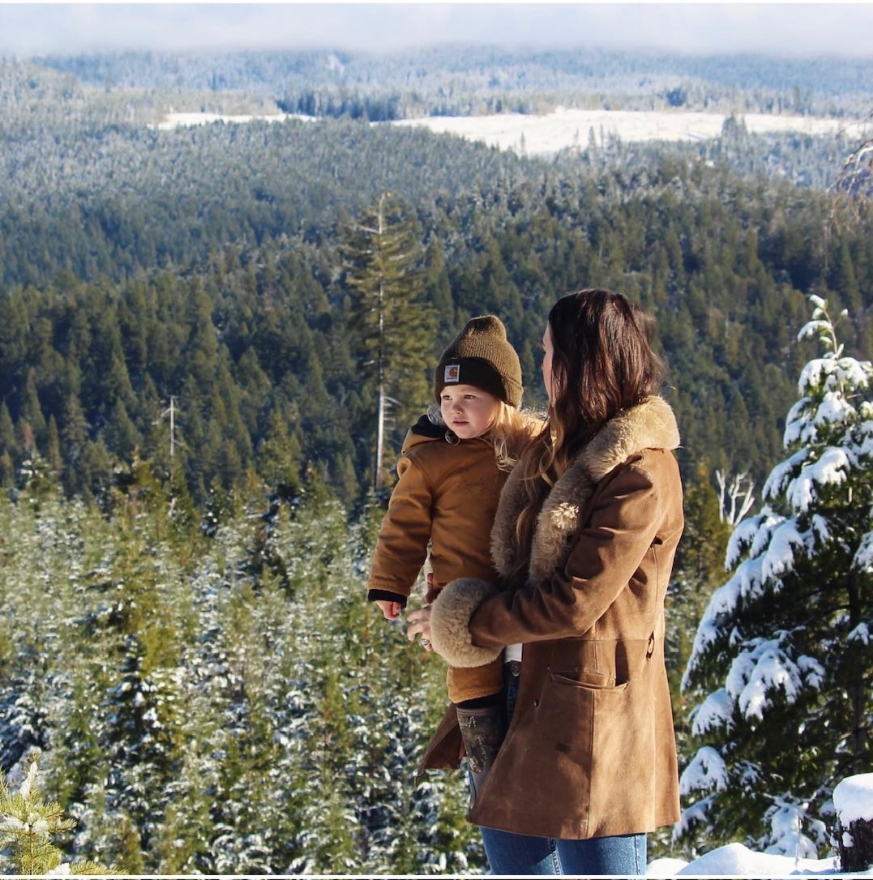 Person holding a child, both in warm coats, overlooking a snowy forest landscape