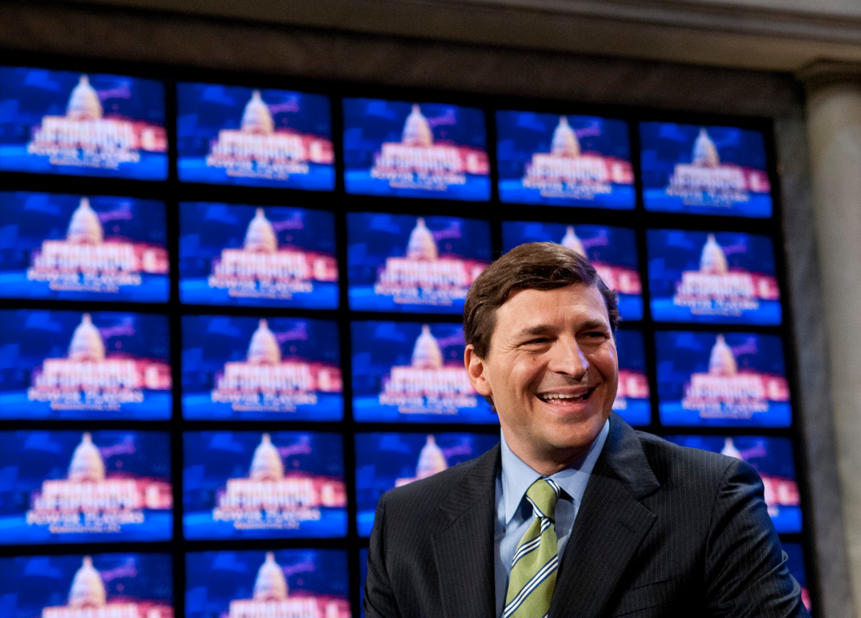 Man in a suit and tie smiling in front of multiple screens displaying a set design resembling the U.S. Capitol