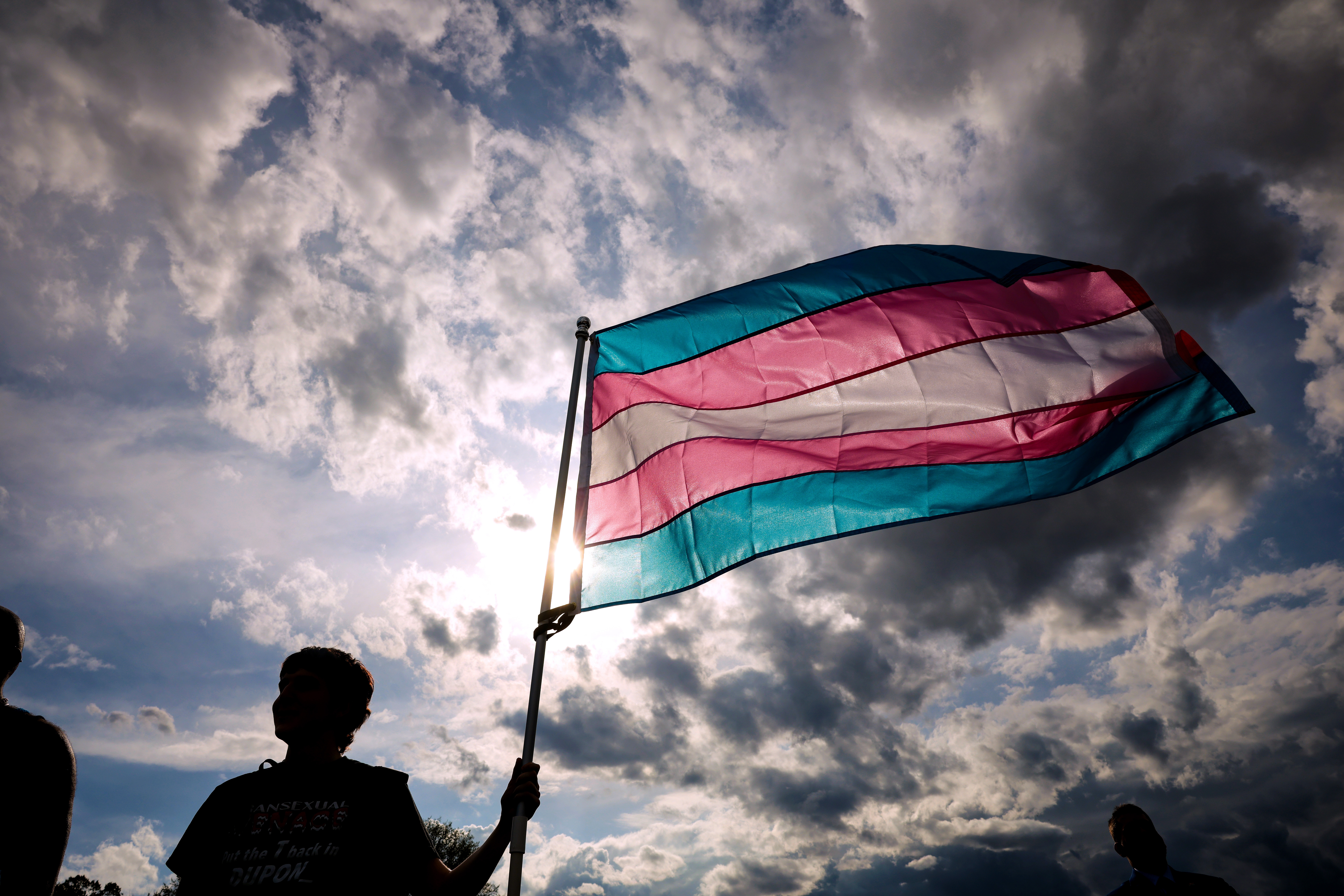 Person holding a transgender pride flag against a cloudy sky, with sunlight streaming through