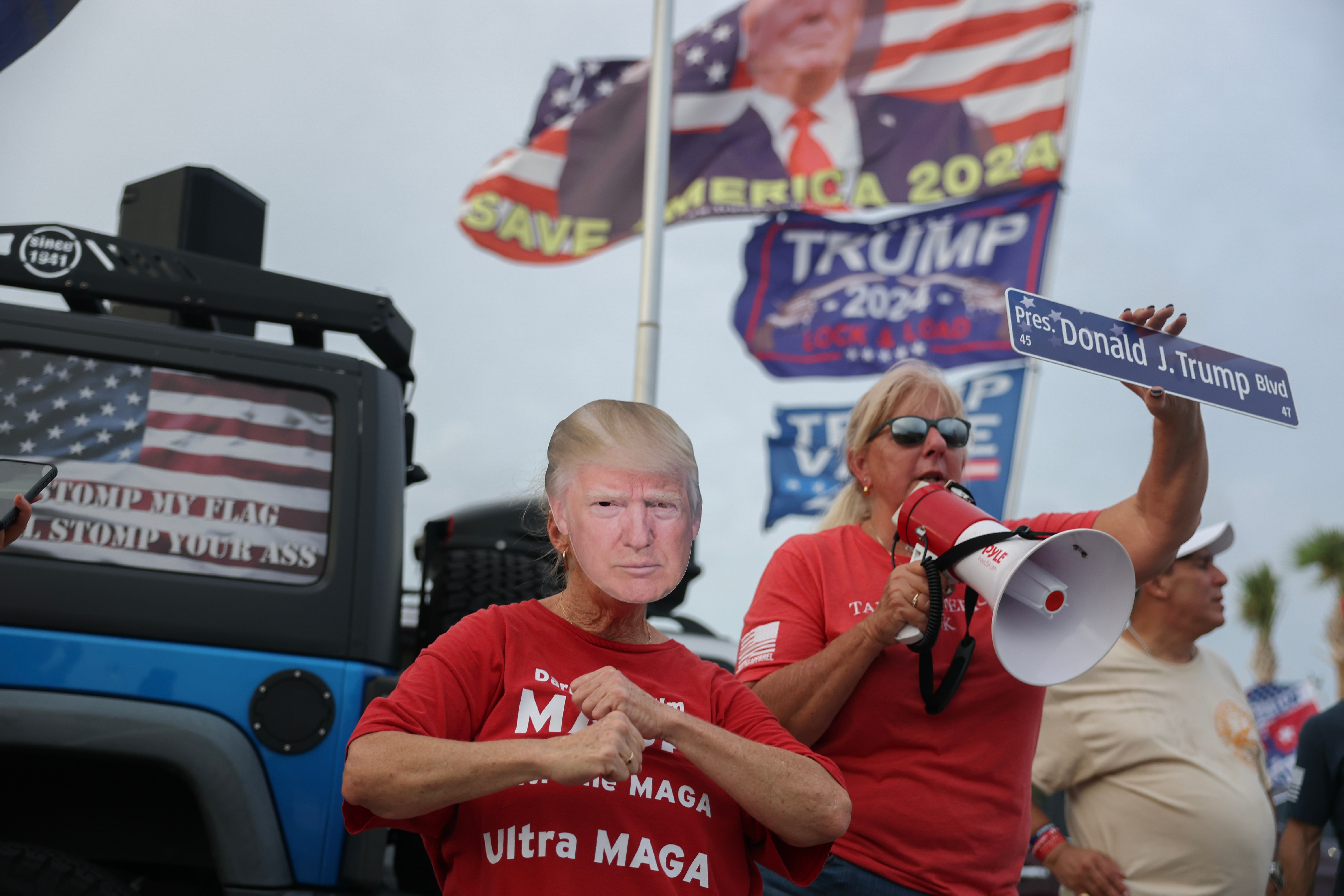 Person in Trump mask at a rally holds a pro-Trump sign, while a woman with a megaphone stands beside