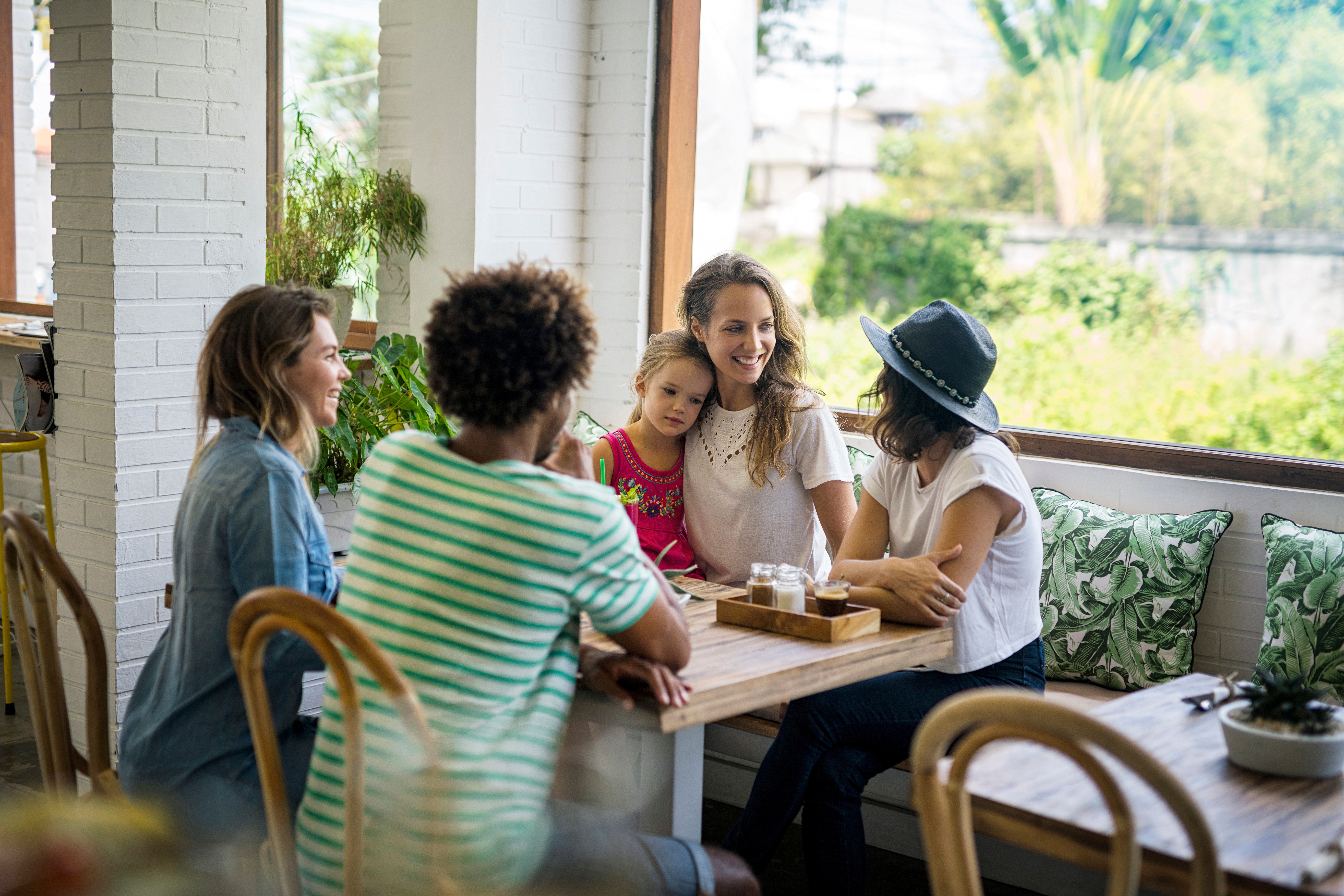 A group of friends with a child at a café, smiling and talking around a wooden table with drinks