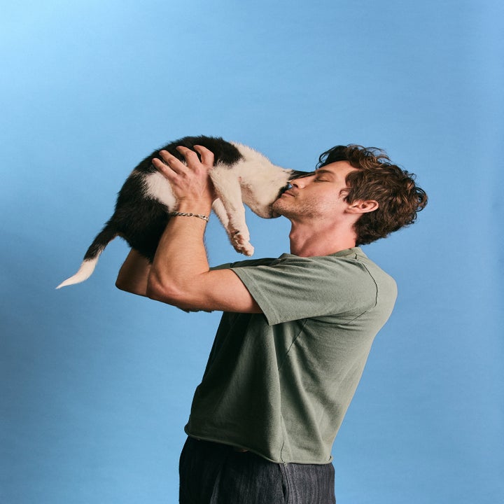 Logan in a casual outfit lifts a black-and-white puppy, bringing it close to their face against a plain background