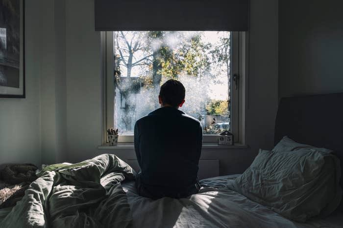 Person sits on bed facing a window, gazing outside at a sunlit view with trees. Room is softly lit, creating a reflective, tranquil atmosphere
