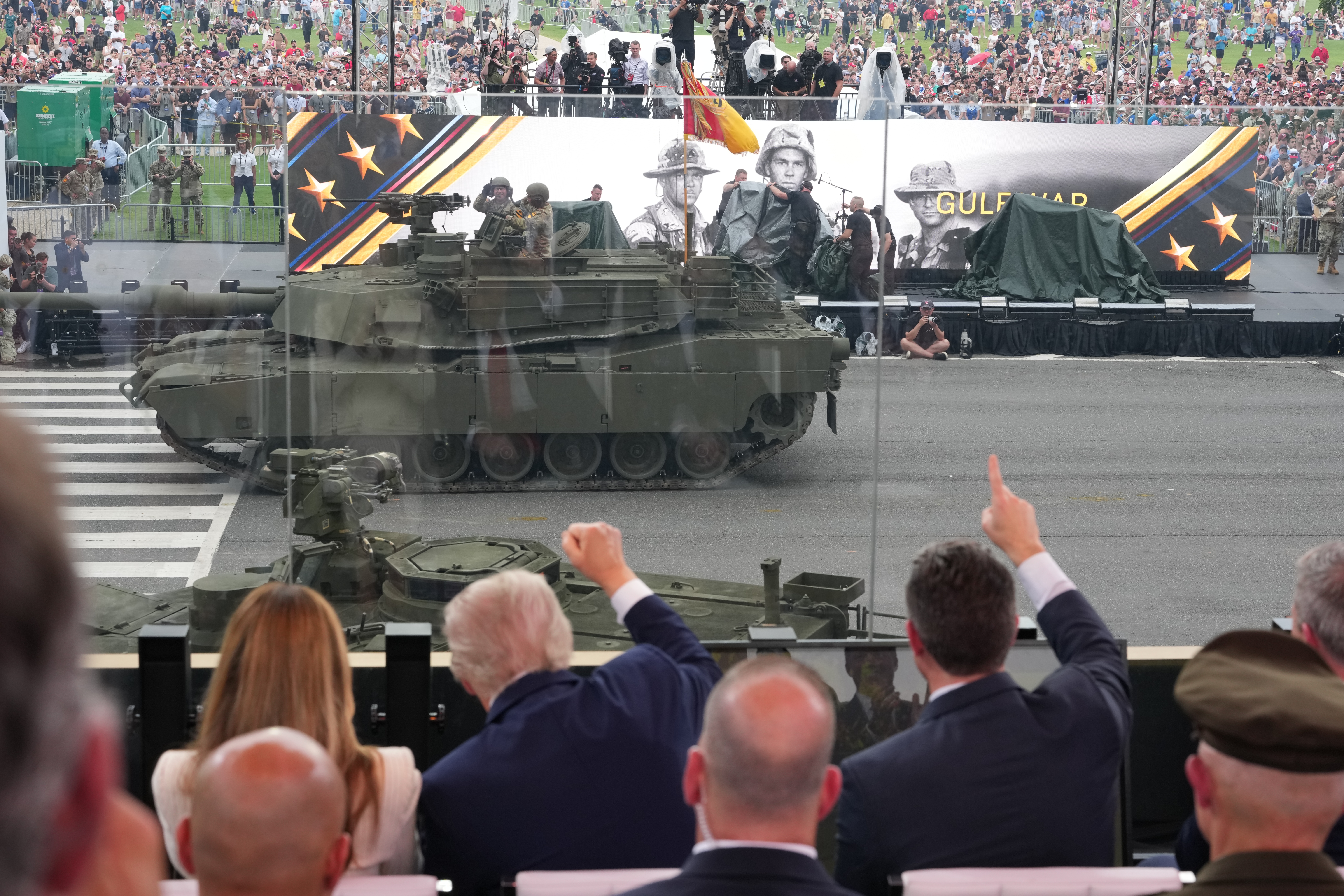 Audience watches military parade with tanks; large Gulf War banner displayed