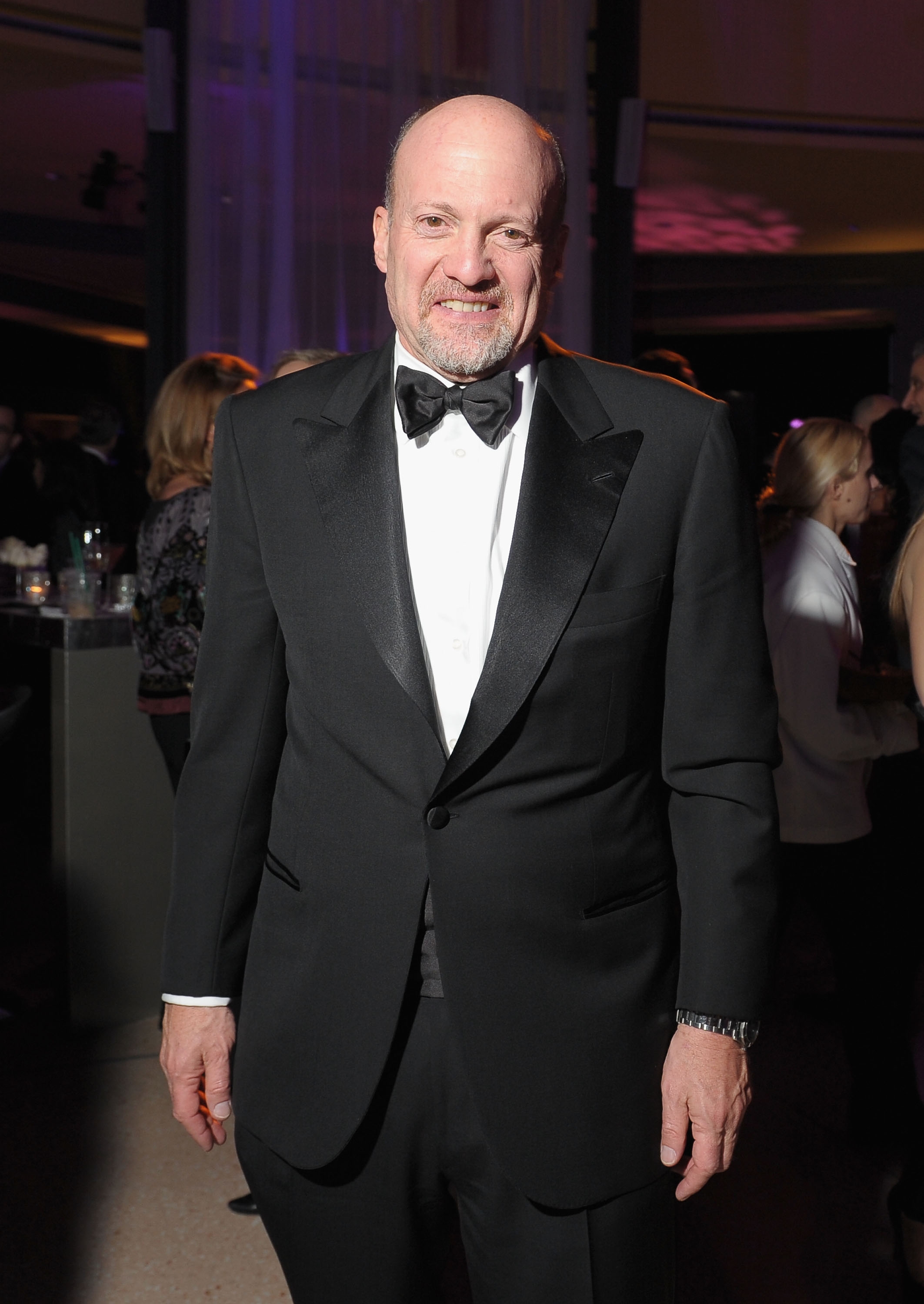 Person in a tuxedo at an event, standing and smiling, surrounded by people in a social setting