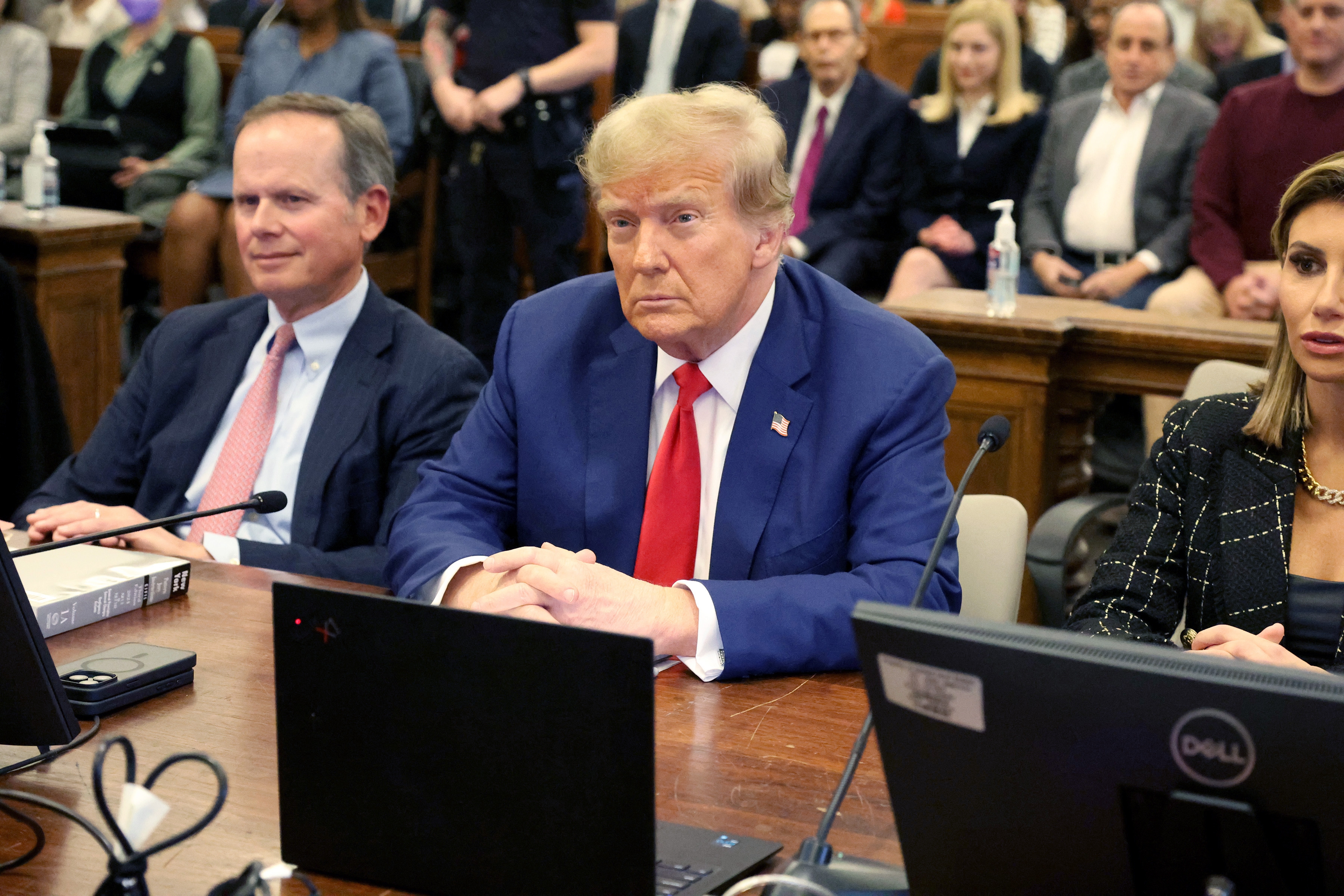 A man in a suit and red tie sits at a courtroom table, flanked by others, during a legal proceeding