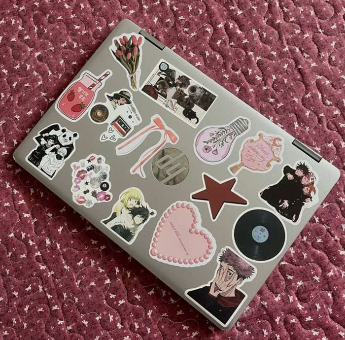 Portátil con varias pegatinas de distintos diseños, incluyendo flores, personajes de anime, una estrella, y un corazón. Fondo texturizado