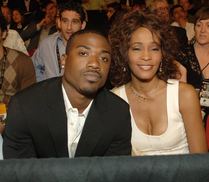 Ray J and Whitney Houston sitting close together, smiling at an event; one in a sleek suit, the other in a sleeveless, elegant dress