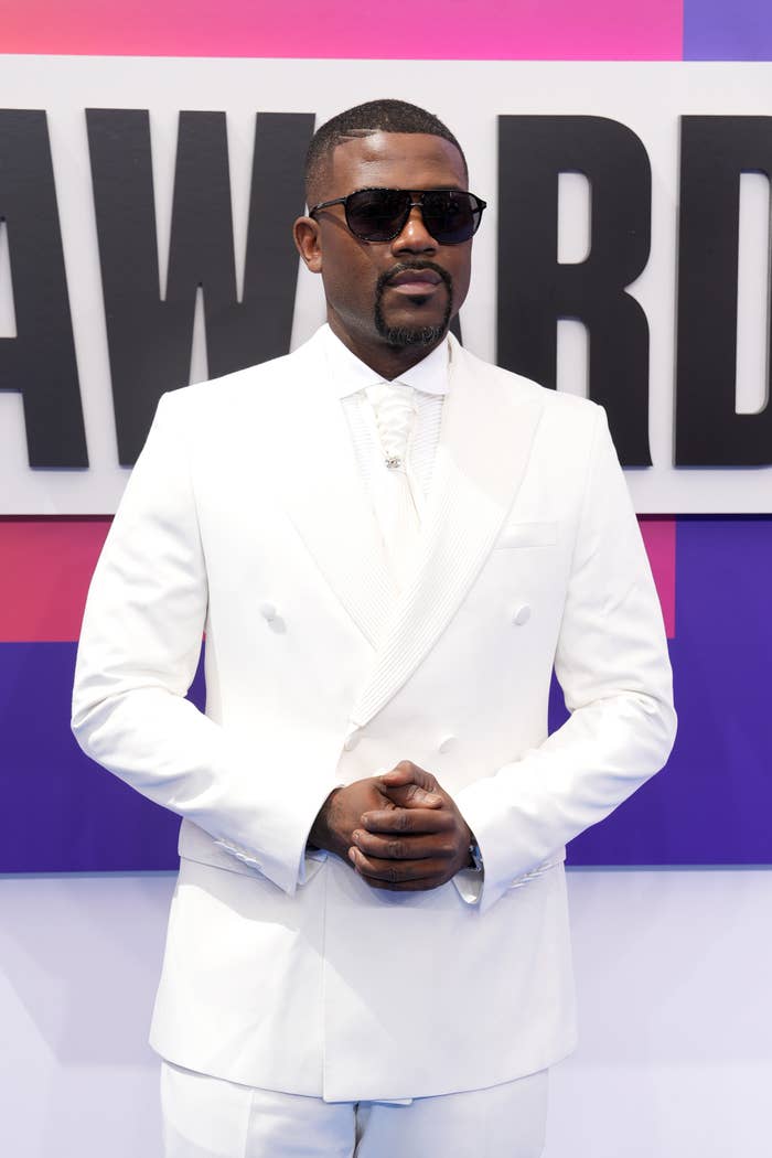 Ray J in a white tuxedo with sunglasses poses confidently in front of an event backdrop