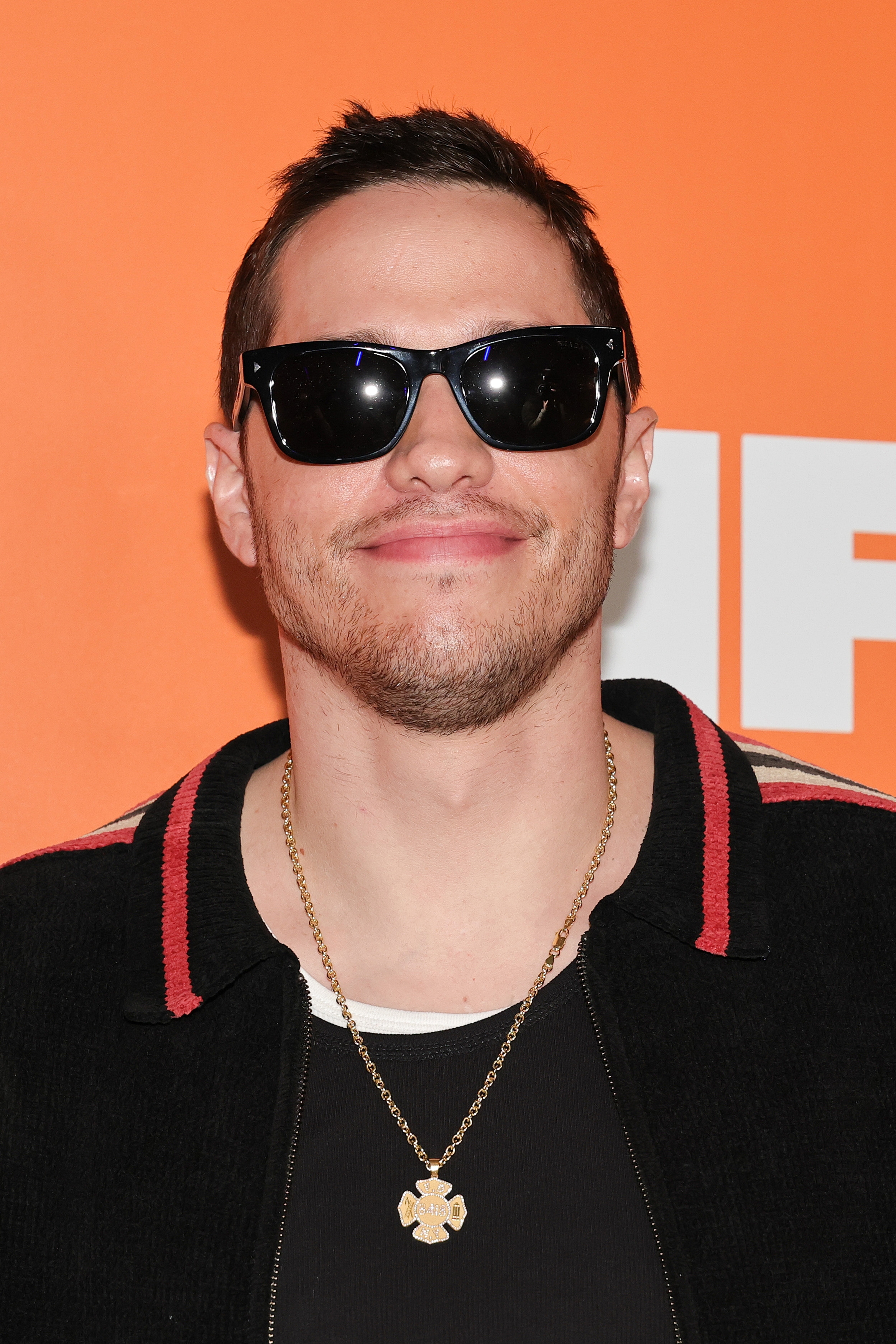 Pete Davidson wearing sunglasses and a necklace, smiling at an event. He is wearing a black jacket with a red-trimmed collar