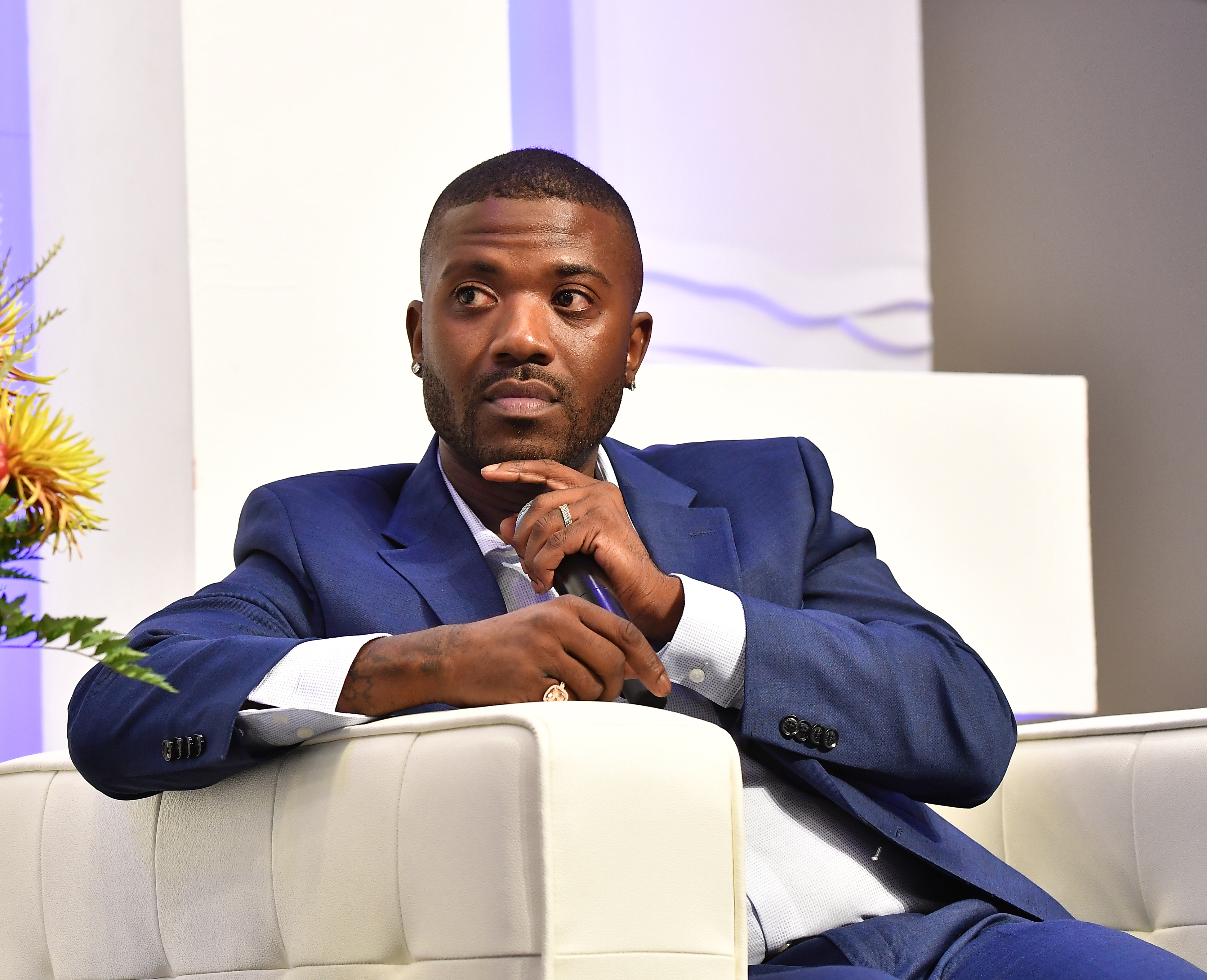 Ray J in a suit sits thoughtfully on a white couch at an event setting