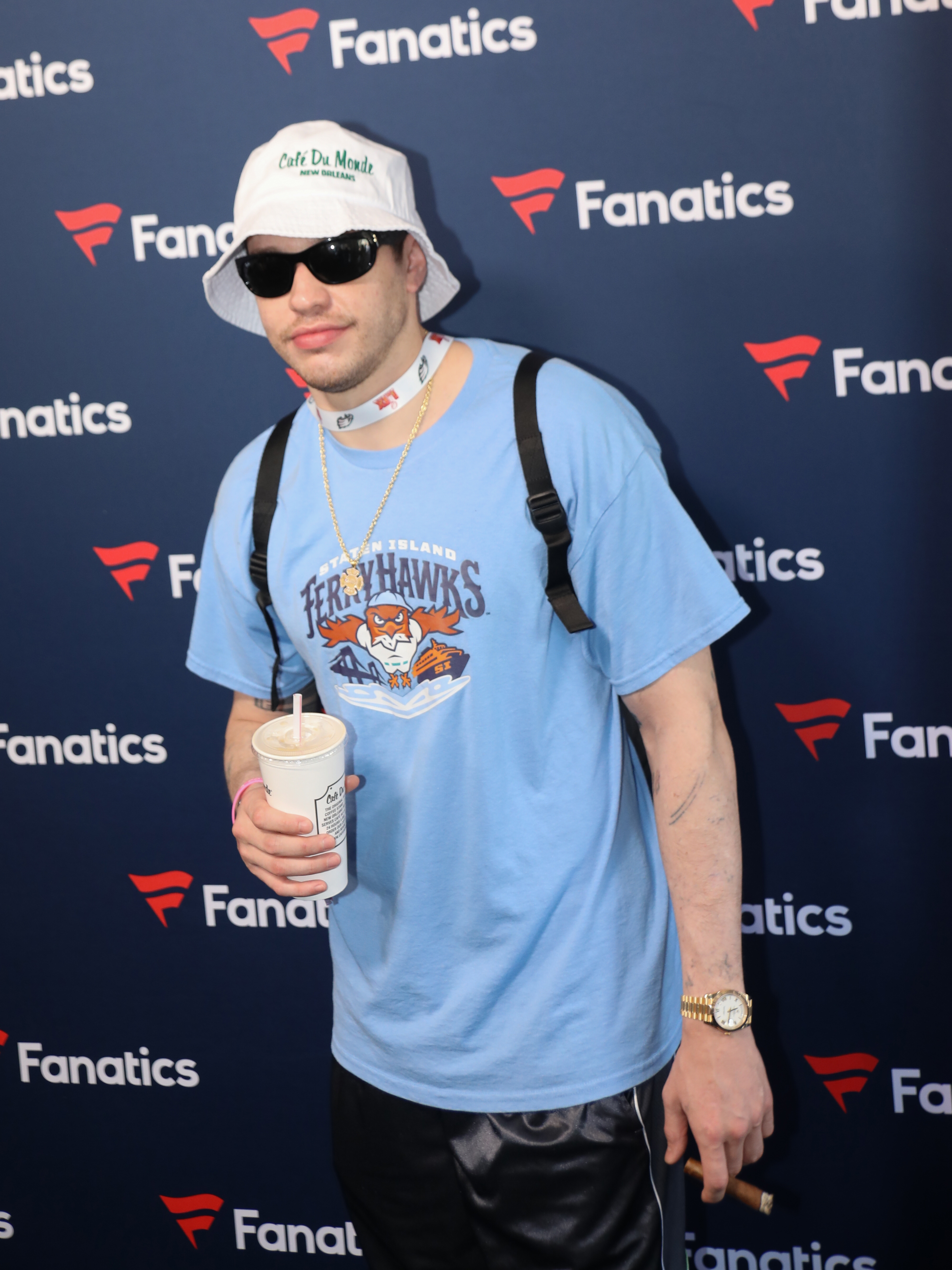 Pete Davidson in casual outfit with a graphic T-shirt, bucket hat, sunglasses, backpack, and holding a drink, poses at Fanatics event backdrop