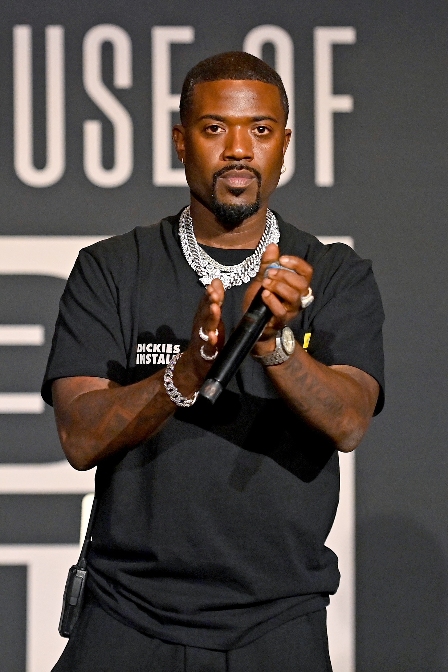 Ray J in a casual T-shirt holds a microphone, clapping hands on stage at an event. Wearing jewelry, he appears to be addressing an audience