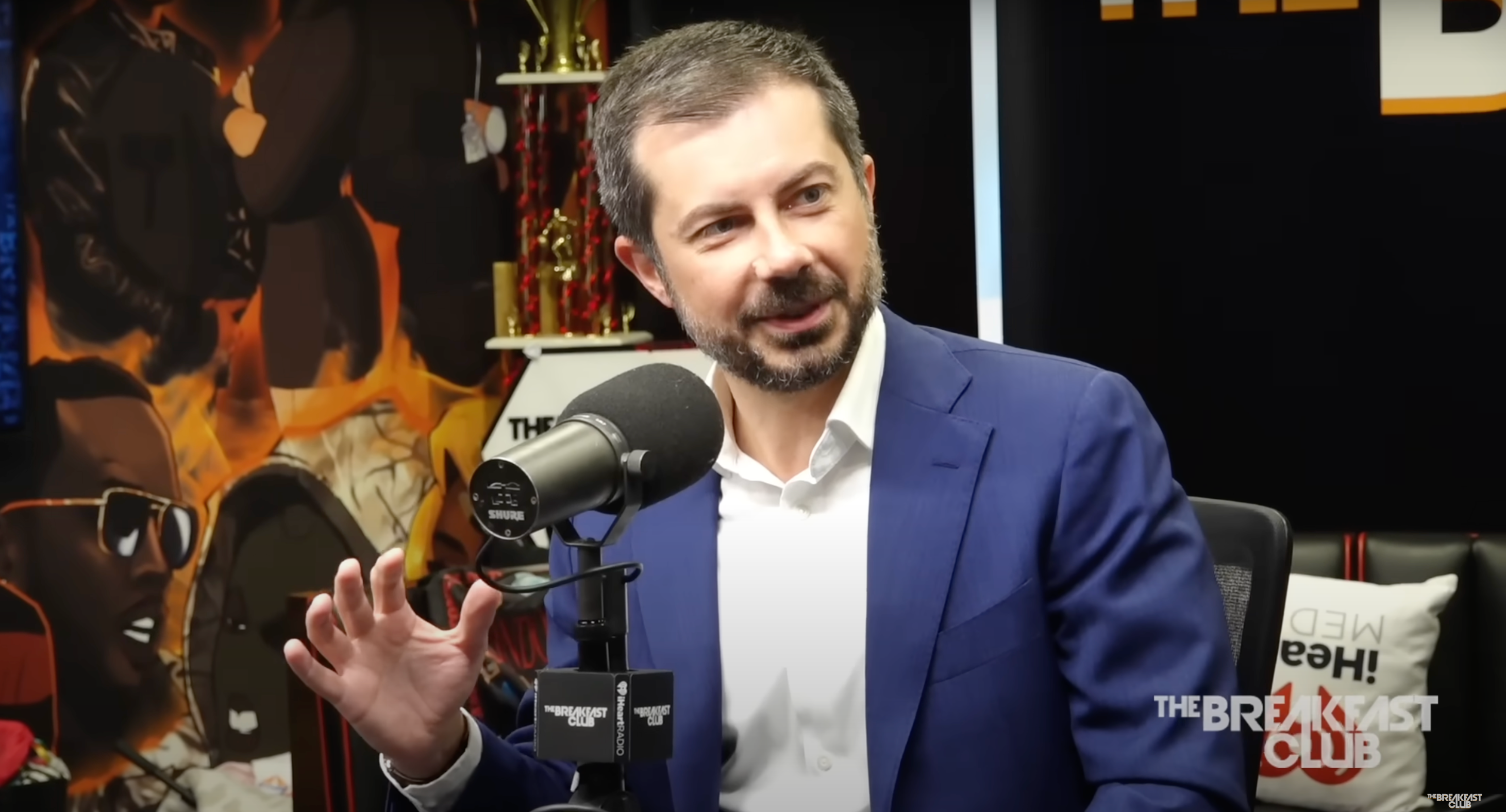A person in a suit speaks into a microphone during a radio interview at The Breakfast Club, sitting in a studio with artwork and trophies in the background
