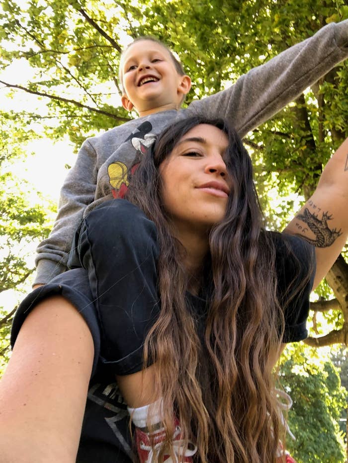 Woman with long hair gives a piggyback ride to a smiling child in a park, both enjoying a playful moment beneath the trees