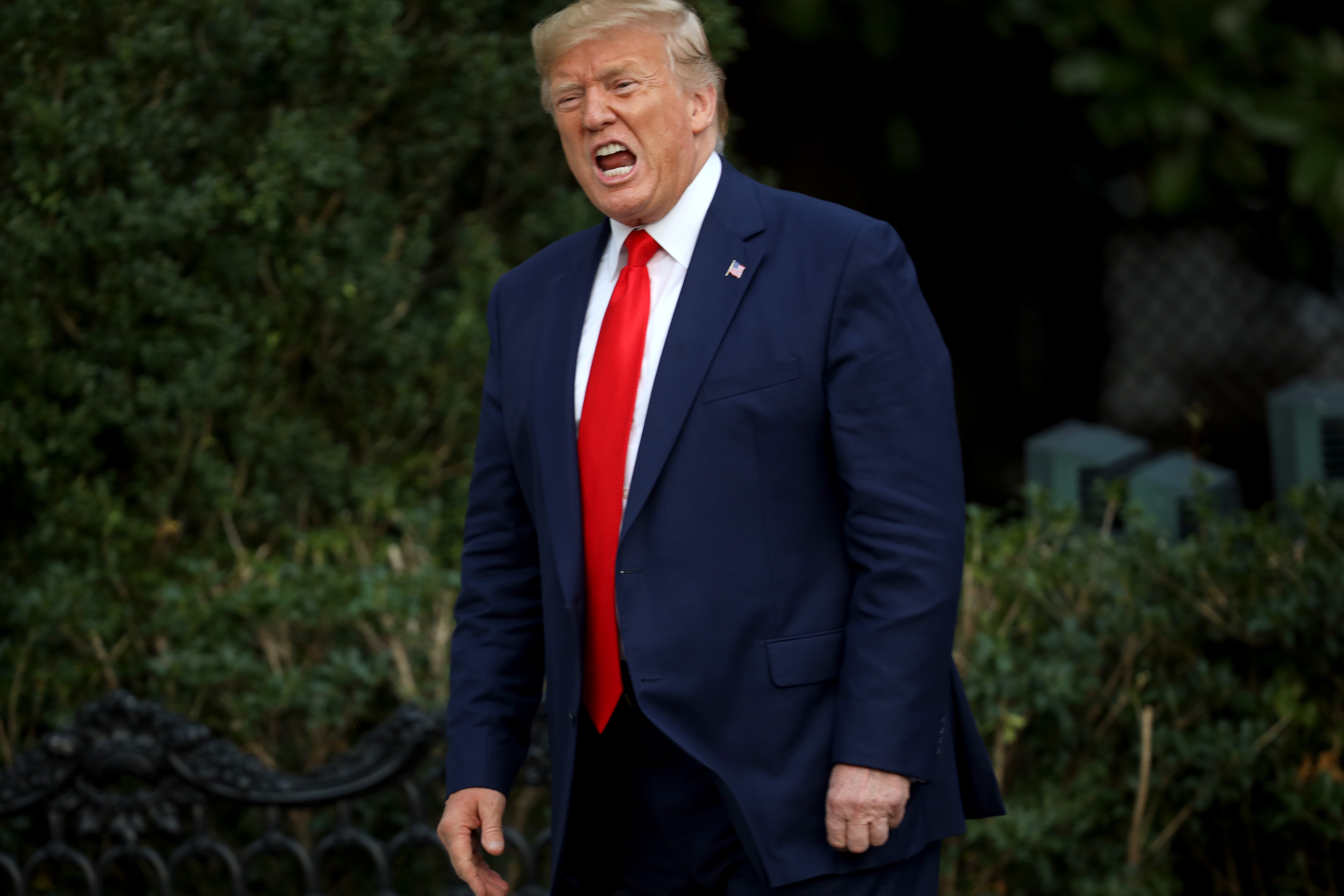 A person in a suit and tie appears to be speaking outside, with greenery in the background