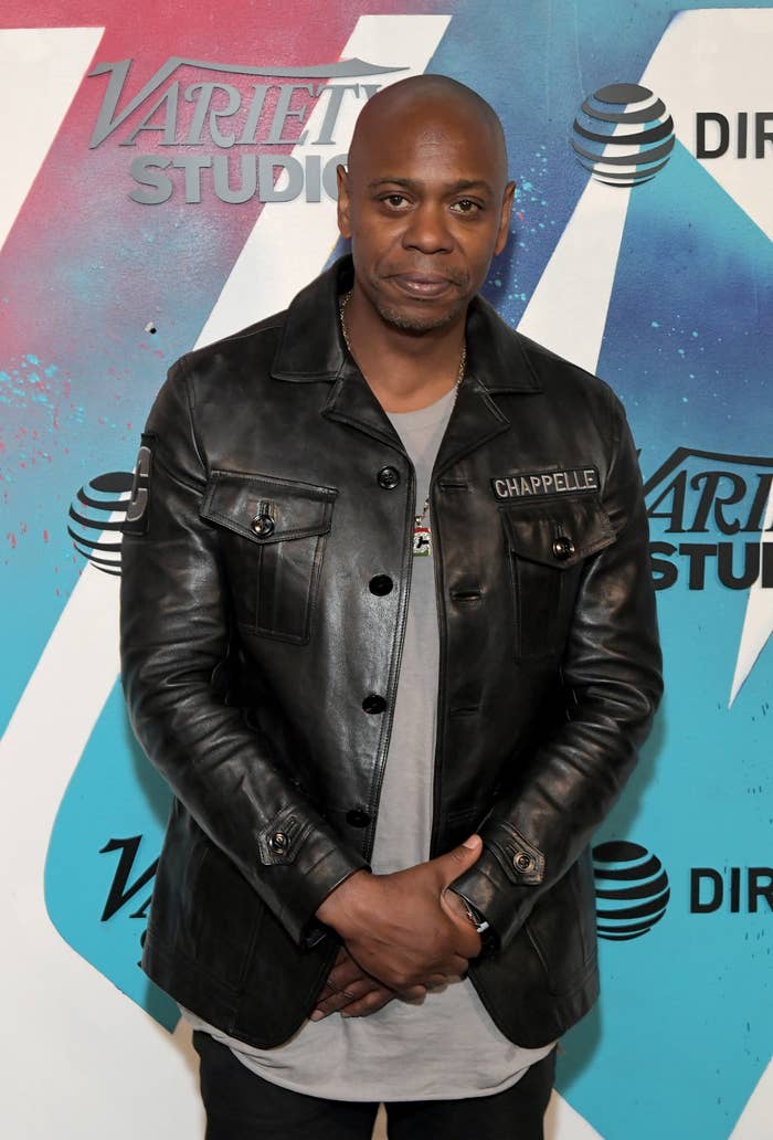 Dave Chappelle in a leather jacket stands at a Variety Studio event backdrop