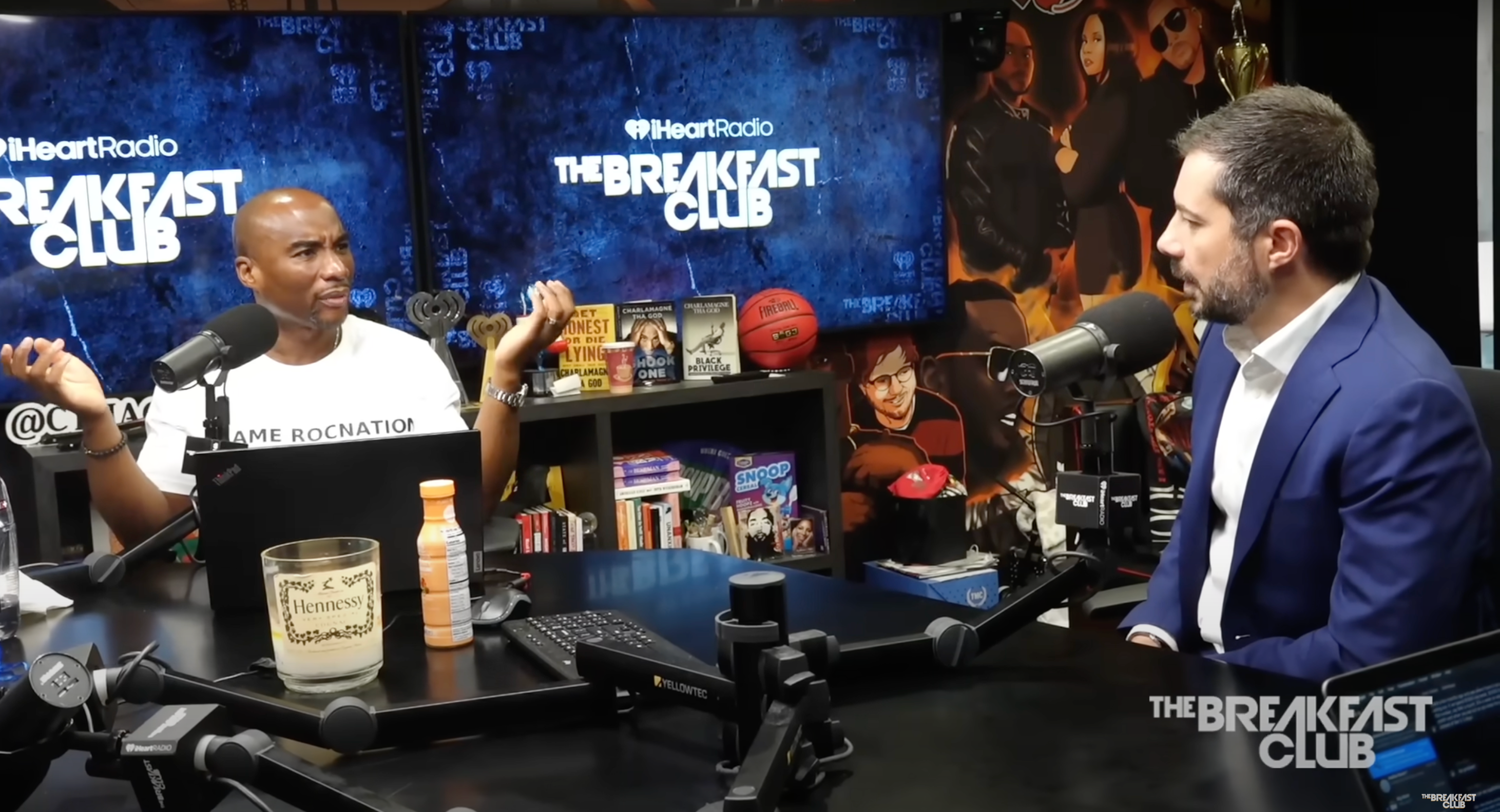 Two people engaged in a conversation in a radio studio, sitting at microphones with &quot;The Breakfast Club&quot; branding visible