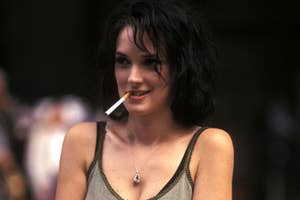 Person with short black hair, wearing a tank top and necklace, holding a cigarette between lips, in a candid outdoor setting